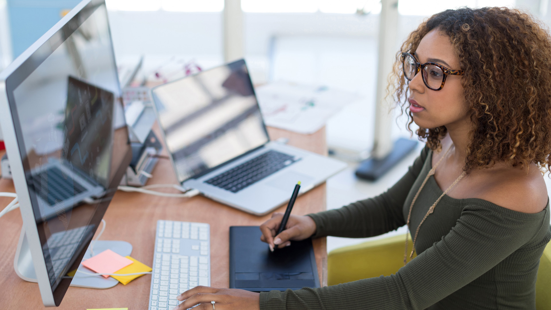 A young woman works at a desktop computer using a stylus on a pad with a laptop nearby.