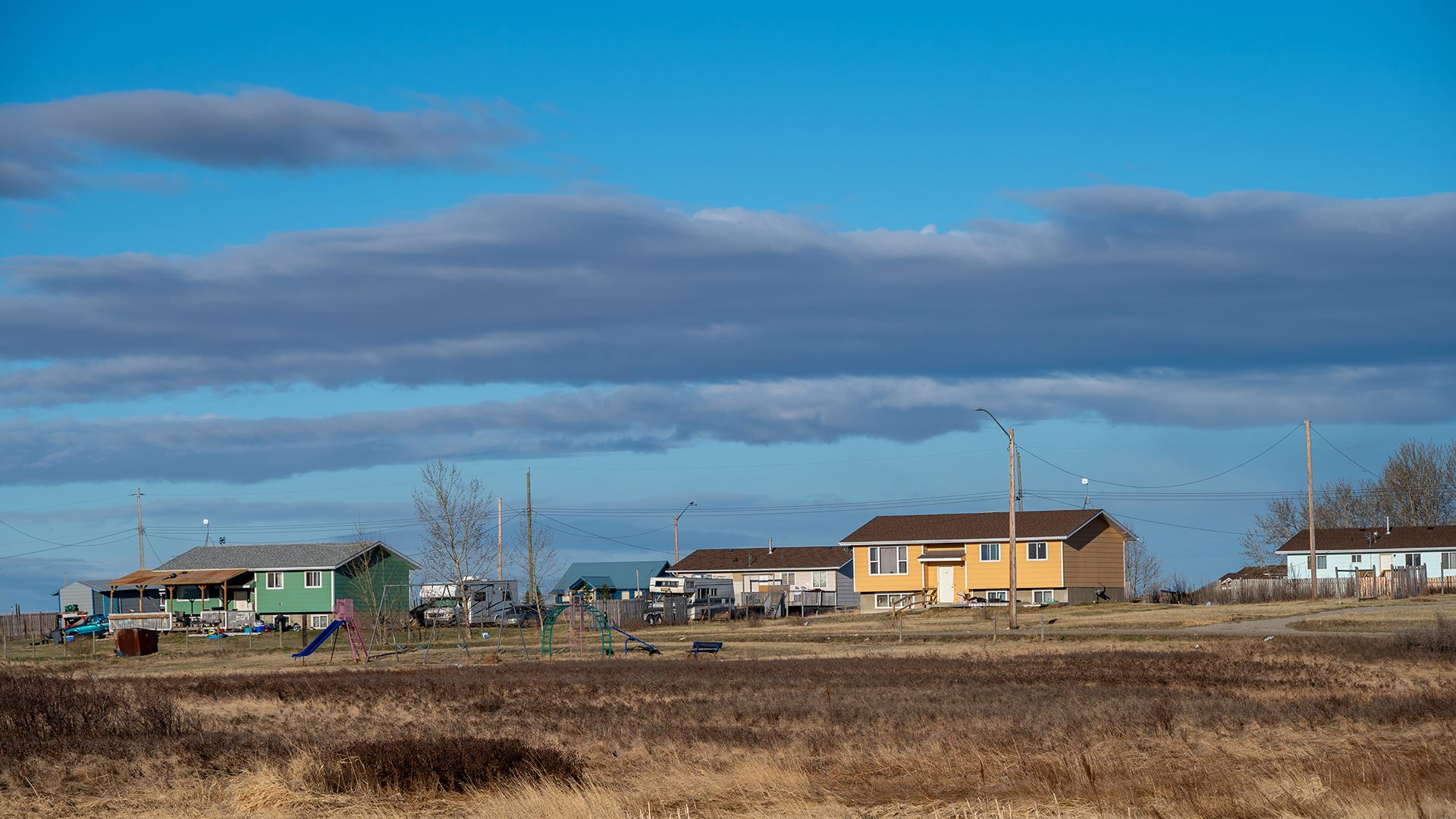 A cluster of bungalows, one orange, one green, sits near a playground among dry brown grass under a big blue sky with long clouds.