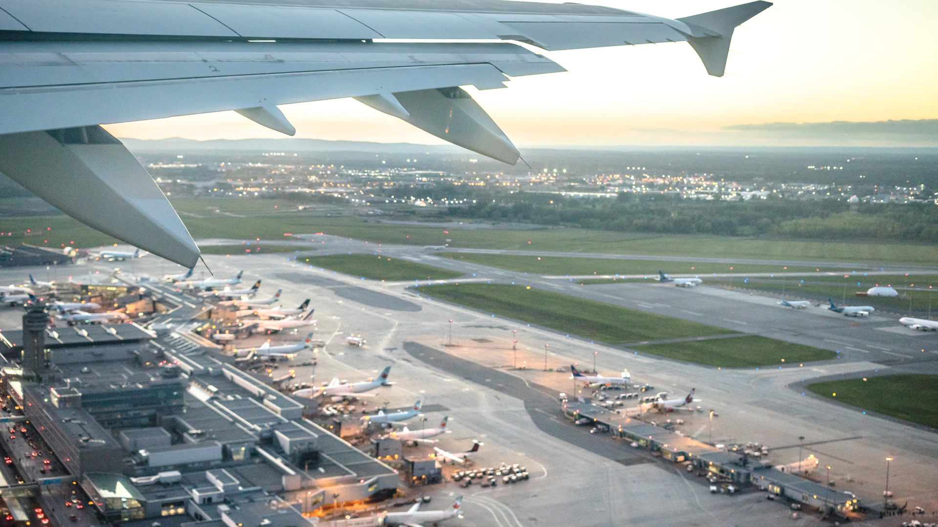 An airplane wing frames the scene of Trudeau airport terminal below, with dozens of aircraft parked at the gates of the terminal.
