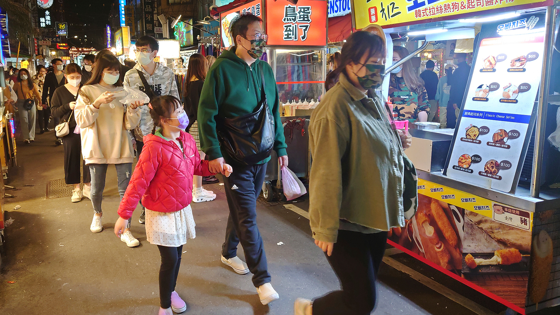 Families and couples walk down an aisle in the market, looking at the food stalls on wheels. Bright signs light up the area.