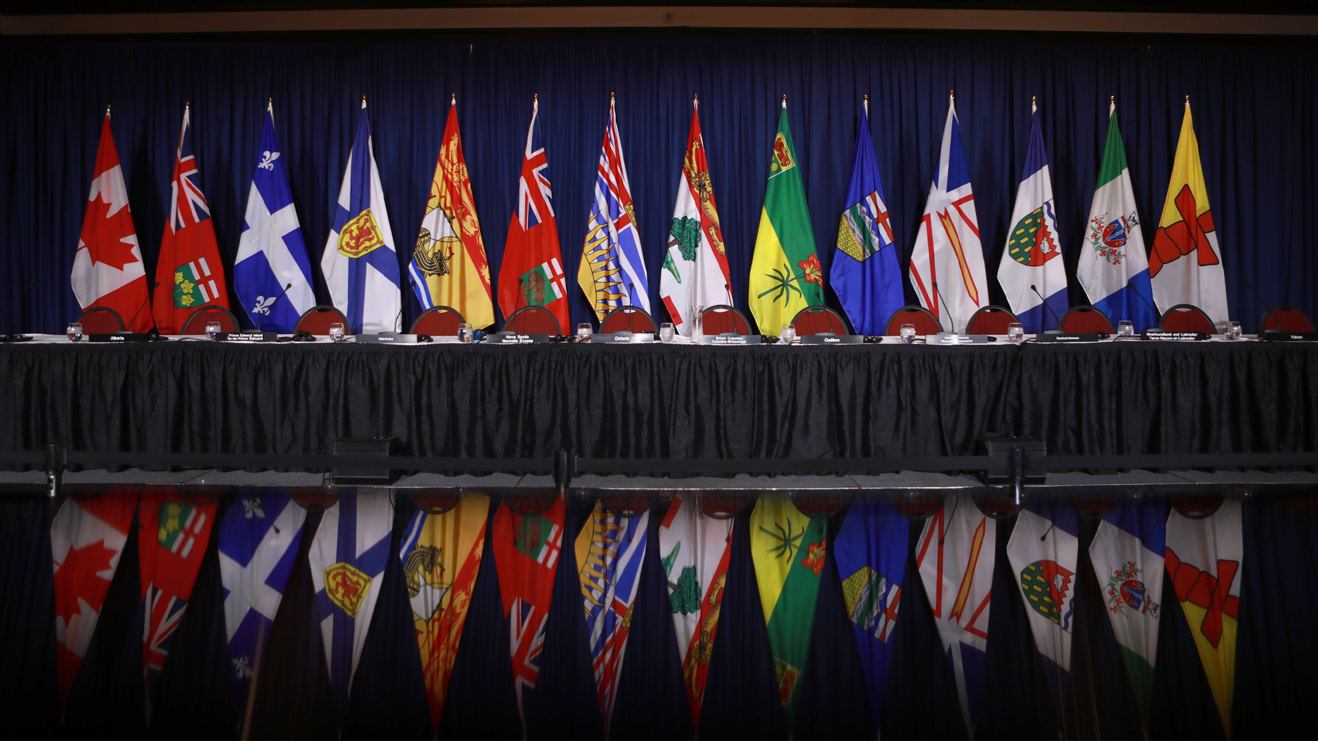 The flags of the provinces and territories are lined up on poles in front of a blue curtain in a conference room.