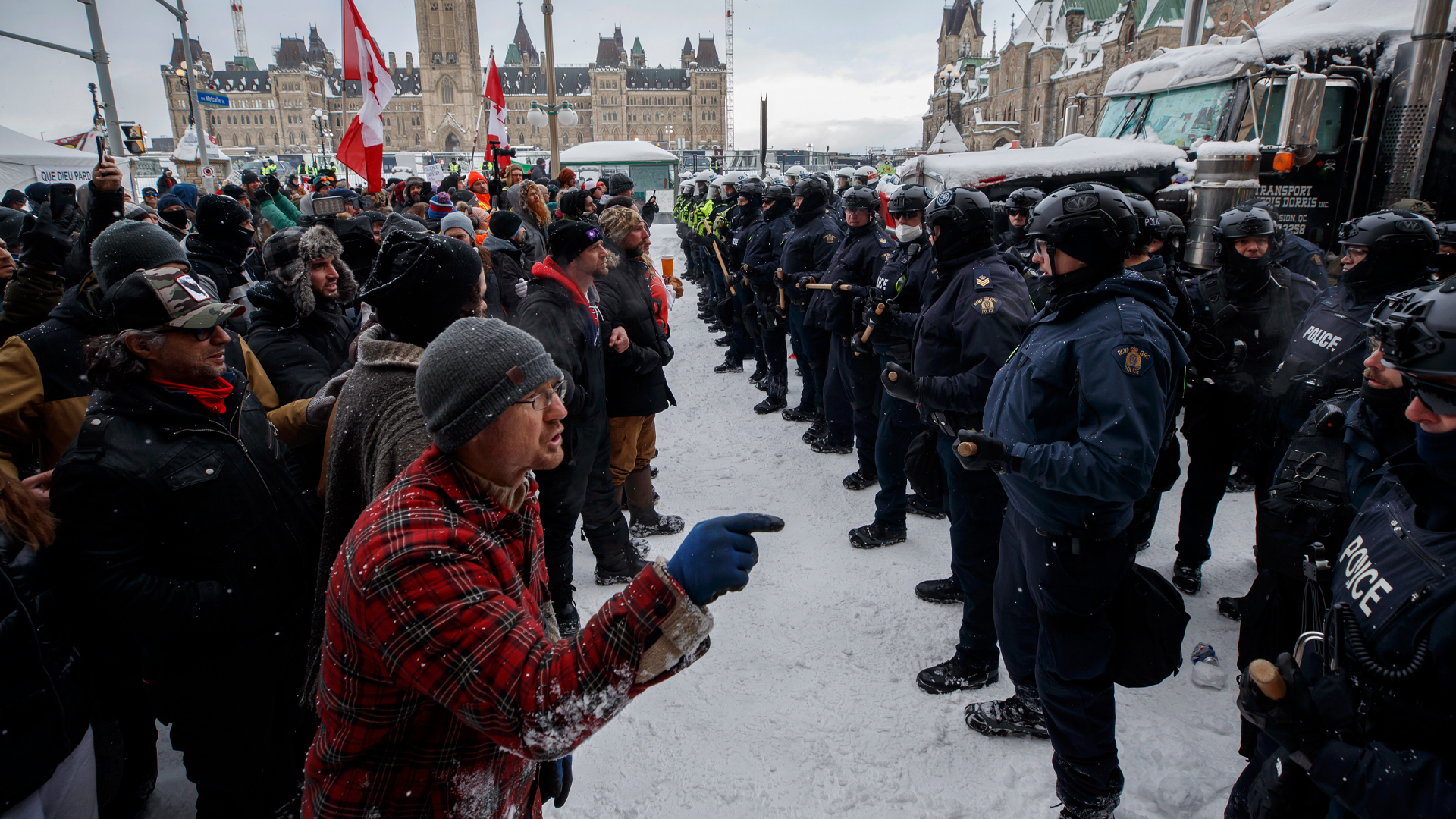 With Parliament Hill in the distance, a large group of people in winter coats, hats and boots, a few holding large Canadian flags, stands opposite a long row of police officers standing opposite them, with a couple feet of space between the groups. One man in the foreground is leaning forward slightly and pointing a gloved finger at the officers opposite him as he says something.