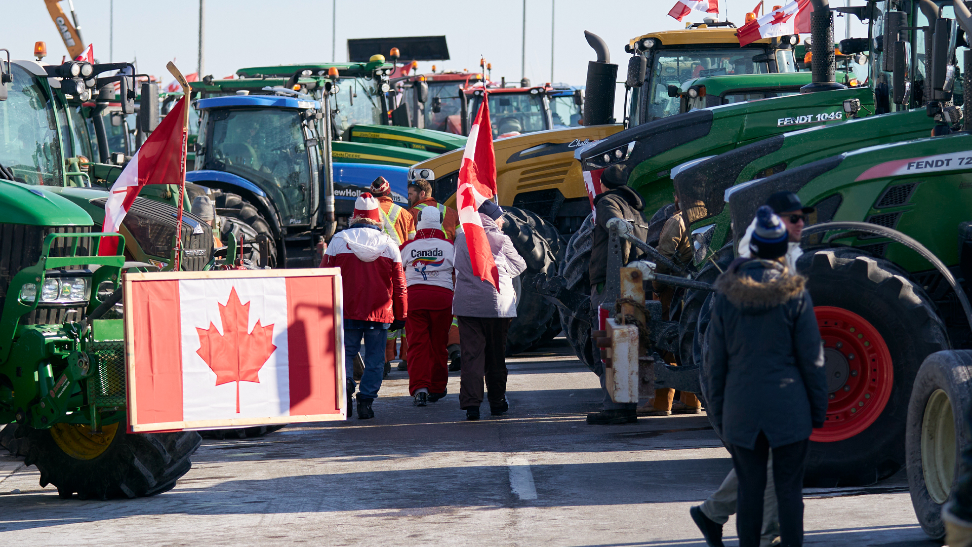 A handful of people in winter jackets and snow pants walk among a dozen parked tractors. Canadian flags are attached to several of the vehicles, and one flag is carried by one person.