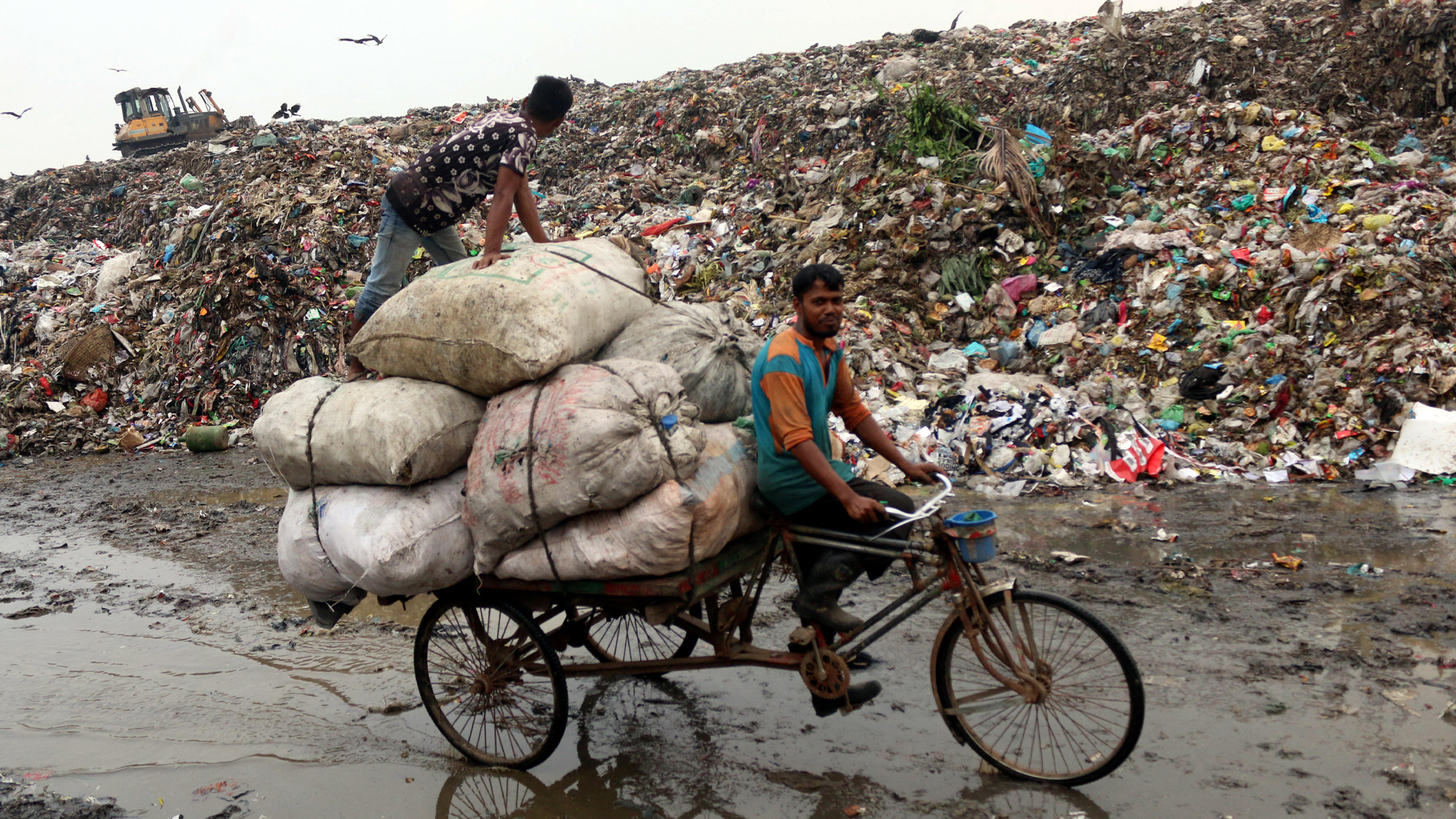 Two men on a bike ride past a mountain of everyday articles piled high, stretching into the distance on a cloudy day. One man is driving the three-wheel bike, and the other is standing on one of eight or so bulging dirty sacks strapped to the back of the bike. Seagulls hover over the dump near a mechanical digger. The ground is covered in a slick of muddy water.
