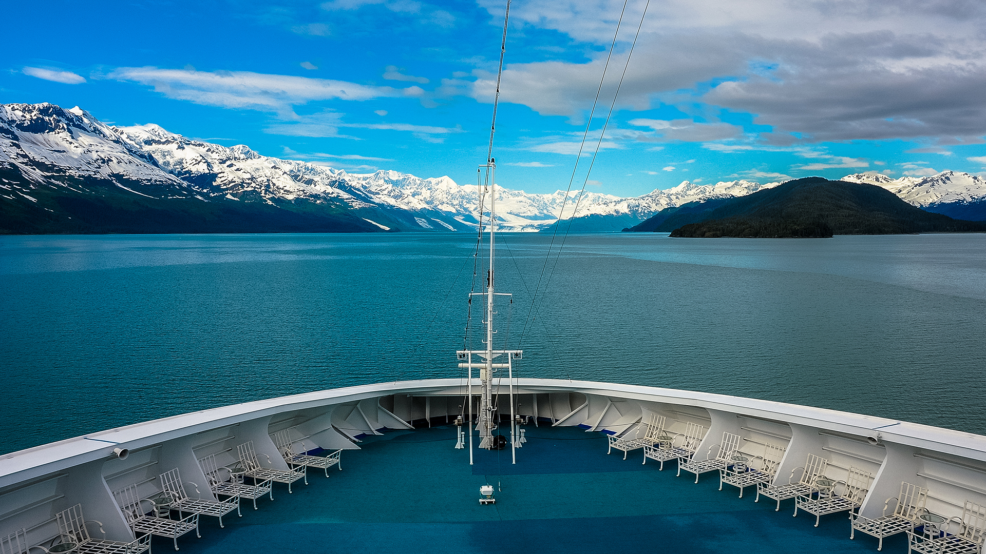 Empty lawn chairs line the deck of a ship at the bow. In the distance is a waterway with various shades of blue ocean and snow-covered mountains with deep blue sky with partial cloud cover.