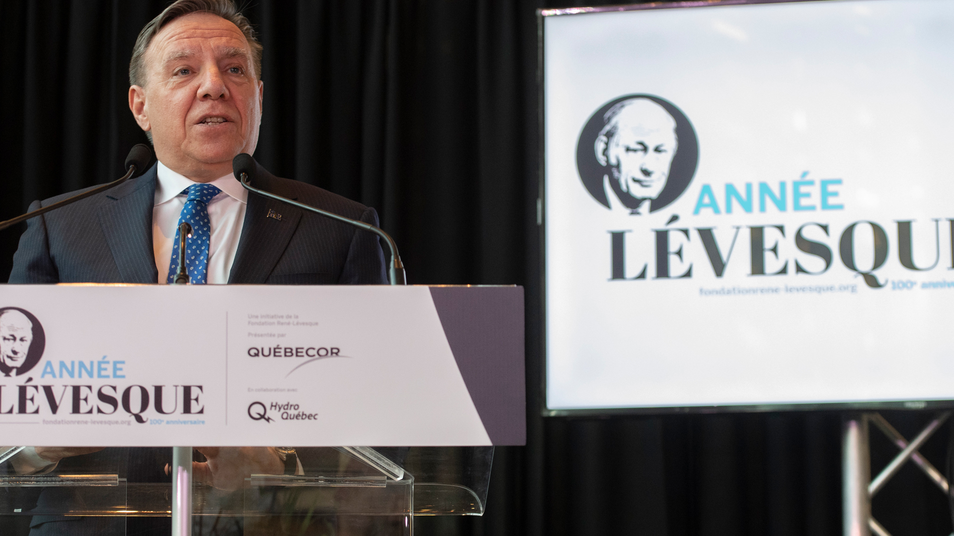 Legault stands at a podium adorned by a sign with a small photo of René Lévesque and the words “Année Lévesque” on it. A larger sign with the same elements is situated behind Legault.