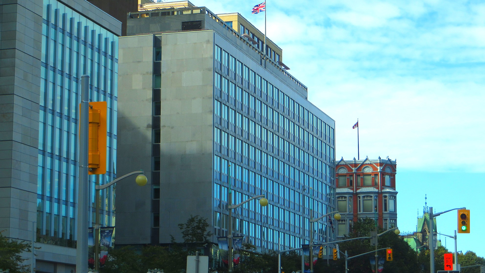 Street intersection view of several buildings: the modern eight-storey building with full-height windows and a shorter, more traditionally elegant building with part of the Parliament buildings in the distance.