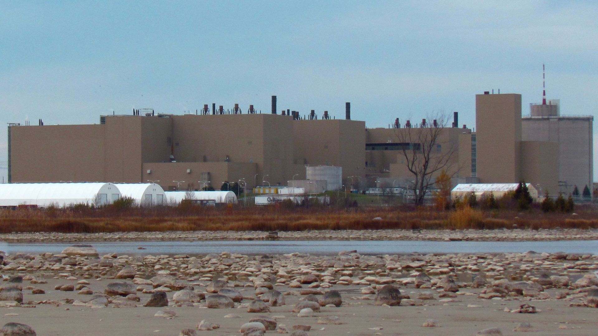 Brown, unadorned, mostly windowless building several stories high, with white dome-covered long structures nearby, sits near the shore of a waterway with sand and small boulders scattered around.