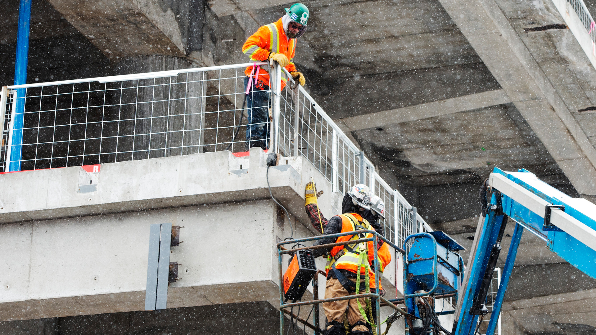 Looking up at man in hardhat and orange safety vest who is standing on a concrete platform with a larger concrete slab above him. He is talking to two similarly clad men who are standing in a small metal cage lifted up by machinery. They are several feet lower than the first man.