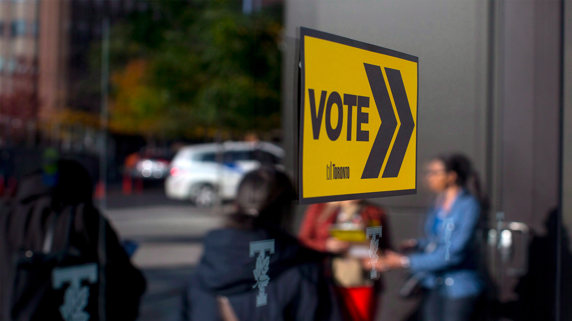 On a pane of glass is a small, affixed sign that says “vote” in bold black letters on a yellow background. Reflected in the glass are four people standing, waiting.