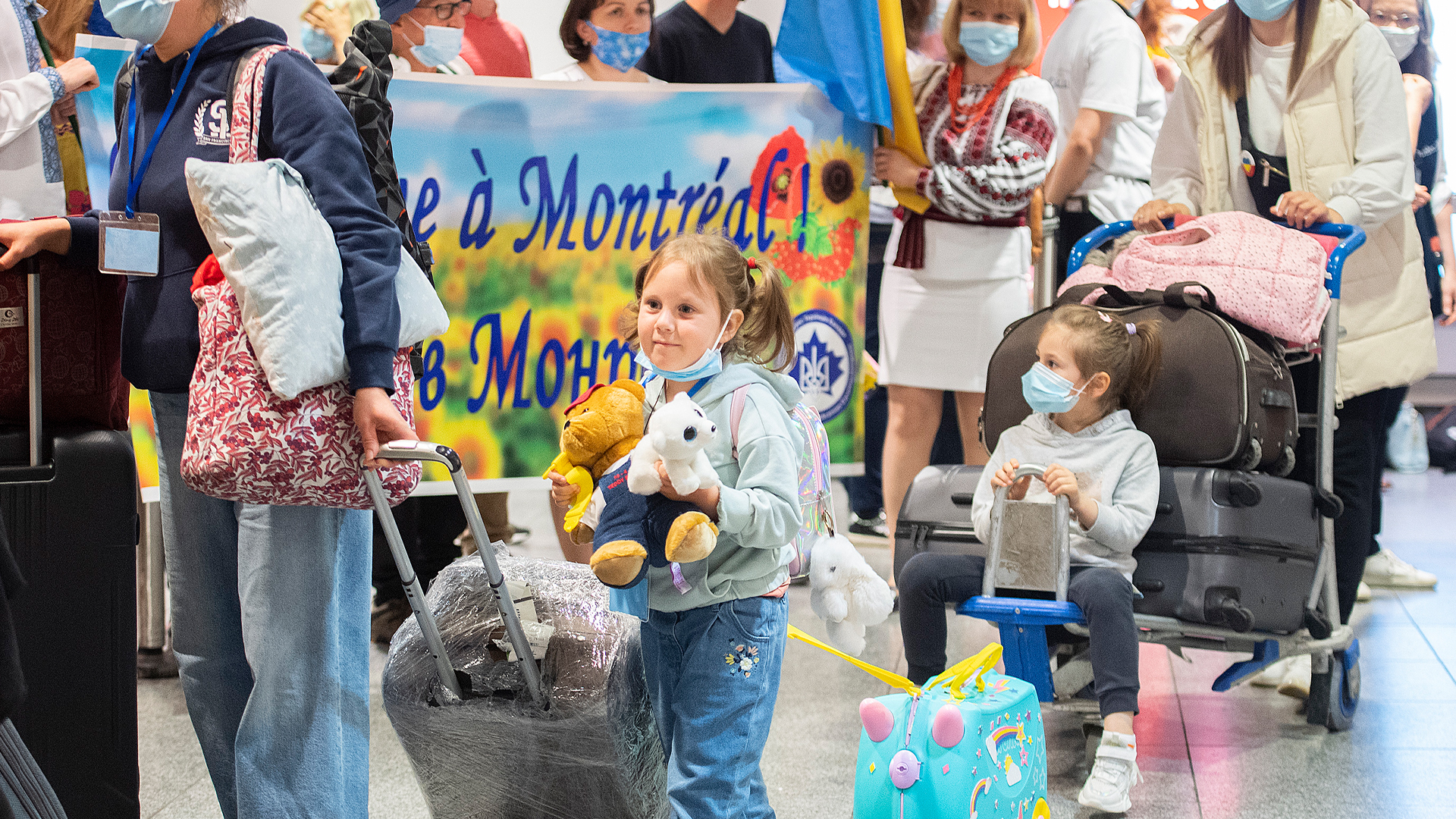 Travellers including a young child holding stuffed animals walk with luggage through an airport arrivals area in front of a welcome sign.