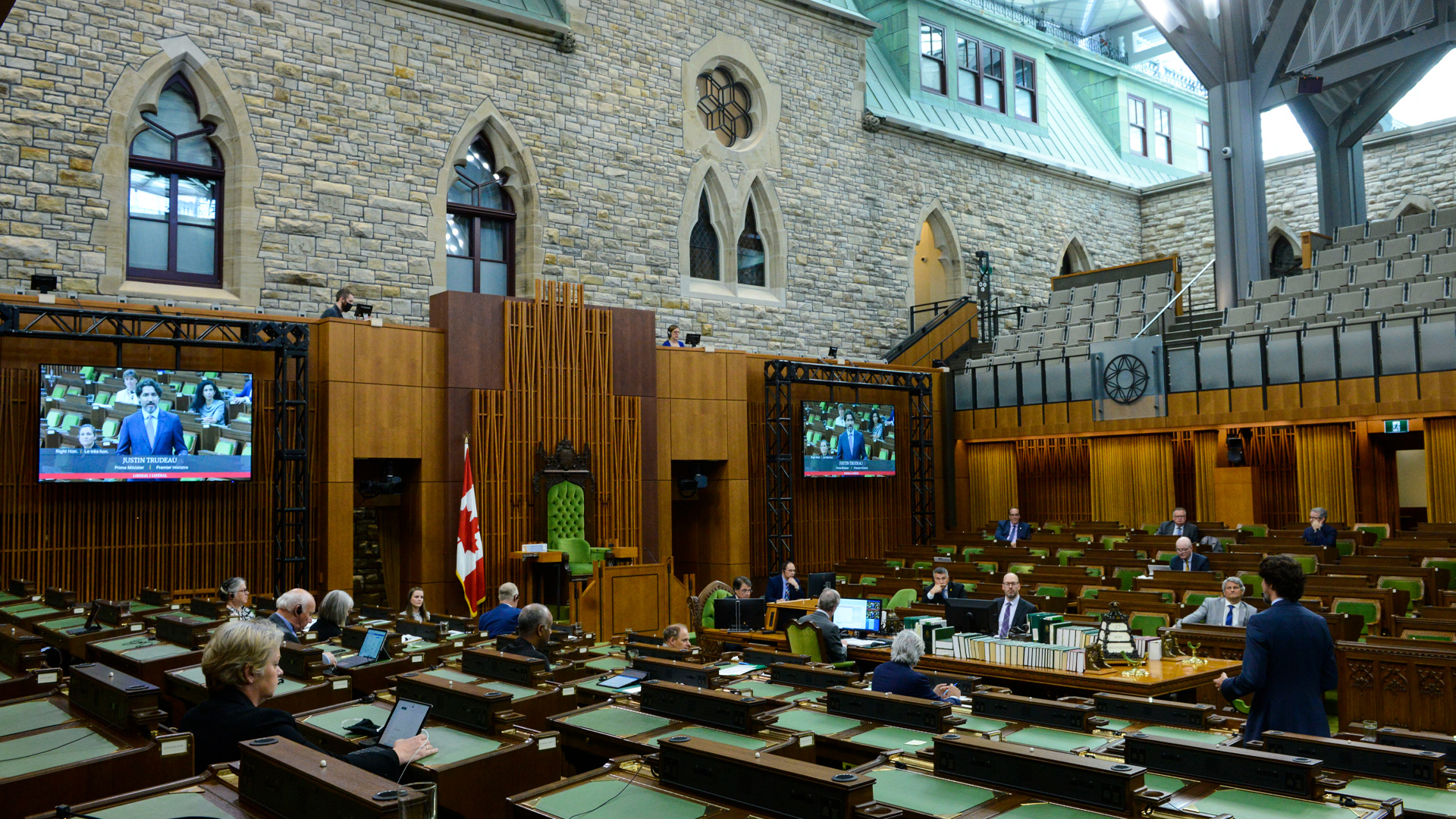 In a light-filled atrium, a handful of men and women sit in the centre of the space, with six rows of desks facing them from either side. The desks are each covered with two side-by-side rectangular green blotters. Seated at only a few of them are a smattering of MPs. The rest are empty. Two large tv screens on one wall broadcast the image of whoever is speaking. In this case, it is the prime minister, Justin Trudeau, who is standing in the middle of the room.
