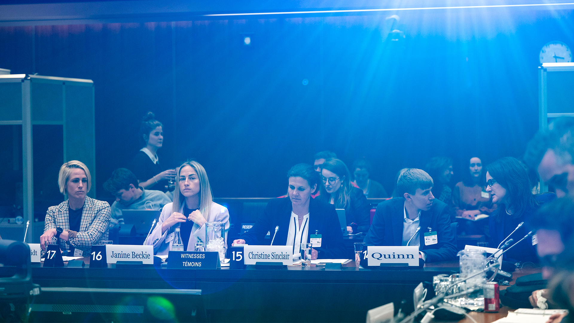 The players are seated at a long table with microphones in front of them, dressed in jackets, with nameplates in front of them. A dozen or so people are seated behind them at two other long tables. A third table of people is nearby. A bright light above casts a blue tint over the windowless room.
