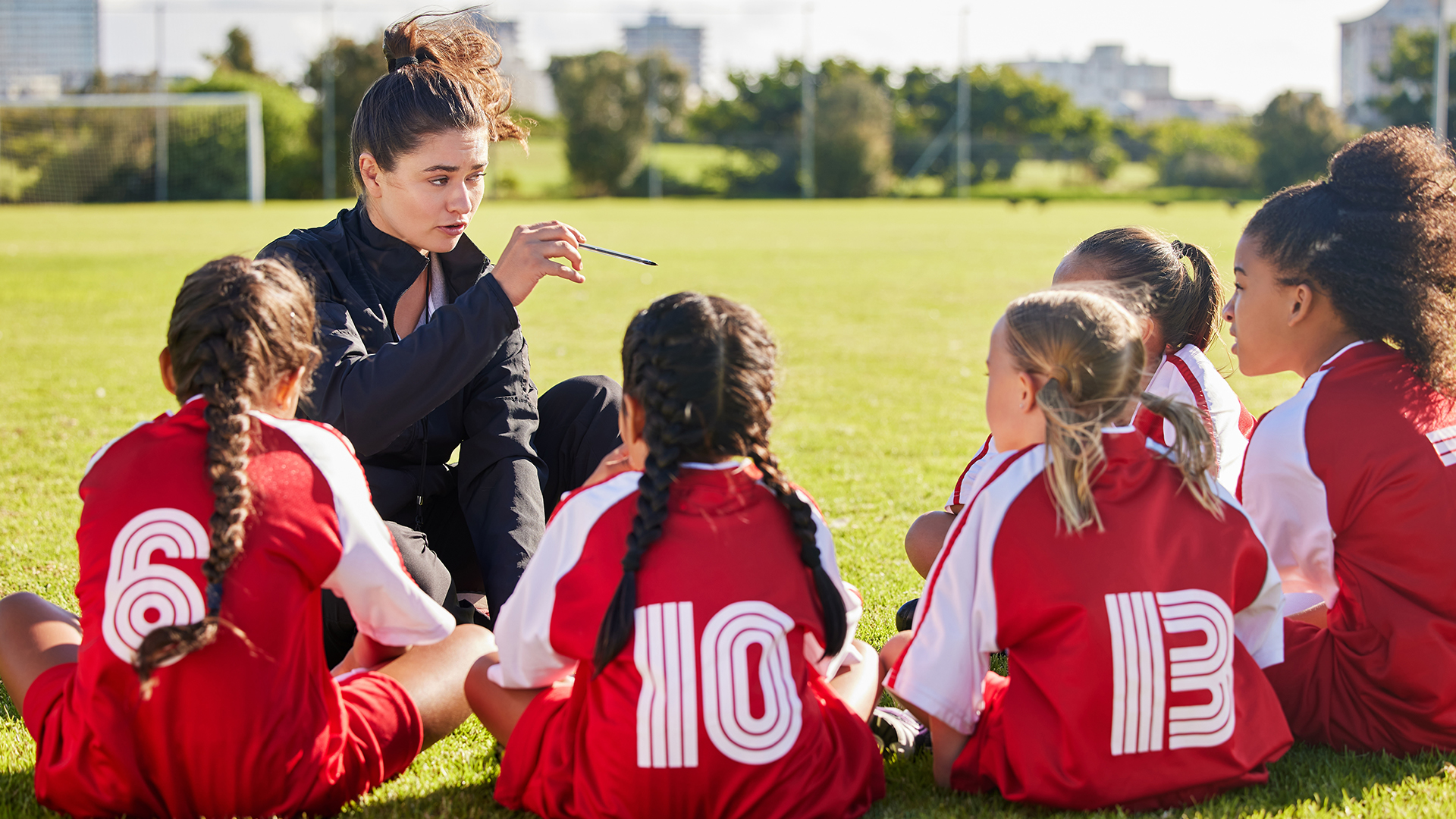 Five adolescent girls are sitting on a soccer field, huddled around a young woman who is telling them something while holding a pen in her hand and pointing it at the girls. The girls are wearing red and white team shorts and loose tops.