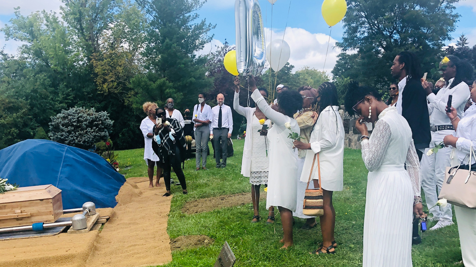 A group of celebrants, mostly Black people dressed in white, stand near a brown wooden casket in a verdant cemetery. Some people are holding balloons while a woman appears to wipe away tears.