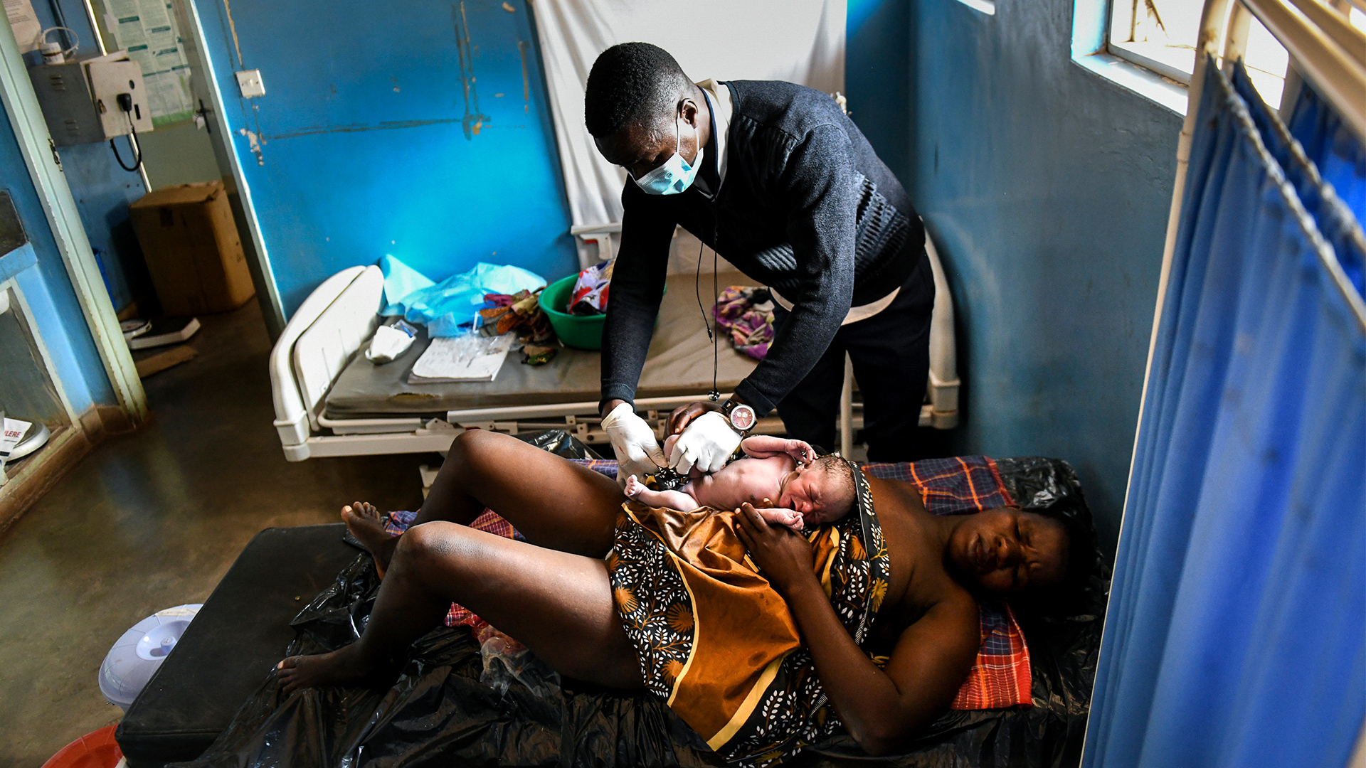 In a small room with blue walls, two simple hospital beds, two small windows, and a white sheet tacked to the wall, a woman lies with her eyes closes, head turned sideways, on a bed with a black mattress, her new baby resting on her tummy. She looks exhausted. A patterned cloth with browns, whites and black covers her midriff, with the baby on top of it. Her knees are up and her legs are bare. The midwife, a man, wears white rubber gloves as leans over to clean the baby.