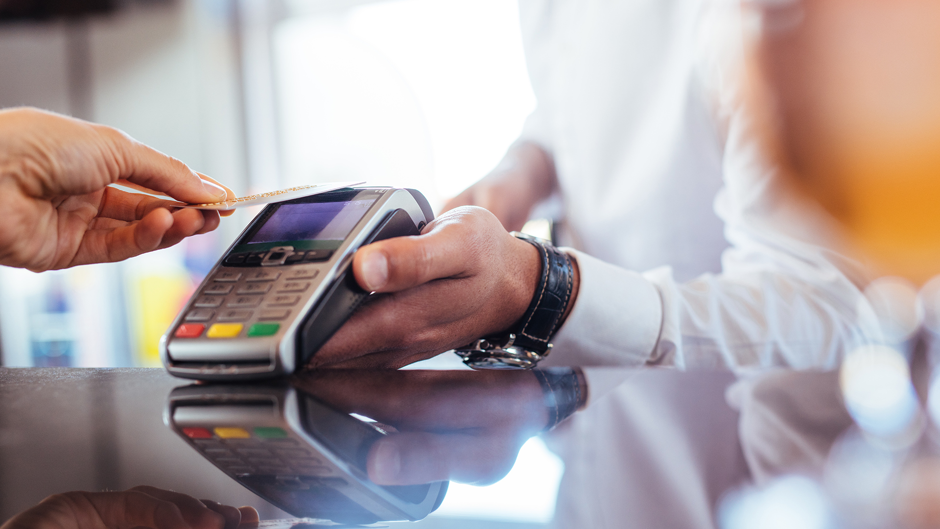 A credit card is tapped on a credit-card payment machine being held by a man wearing a wristwatch with a dark-brown leather strap and a white shirt.