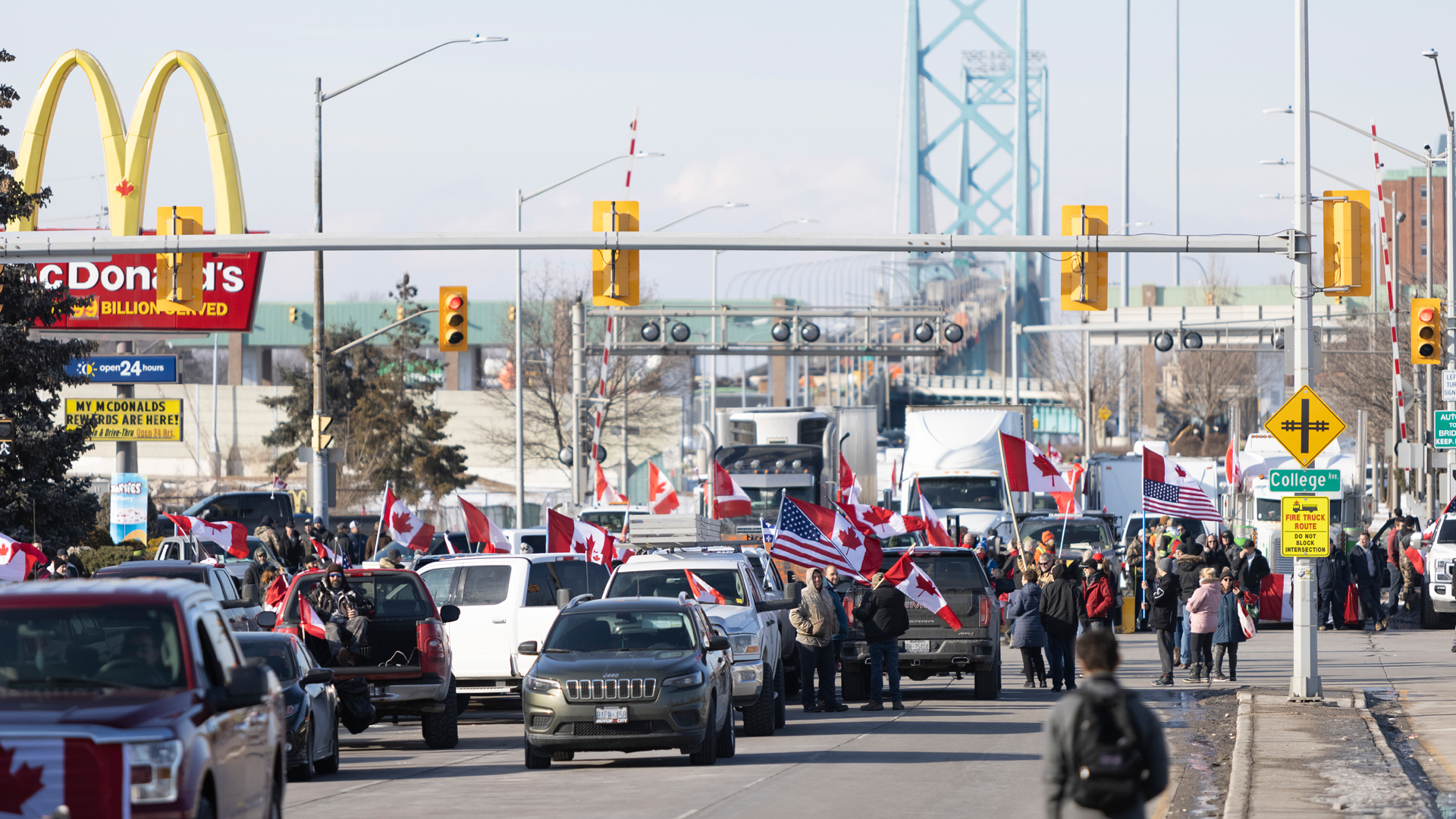Des camions et des voitures sont garés sur les voies menant au pont, que l’on aperçoit à l’arrière-plan. Des groupes de personnes se tiennent debout, brandissant des drapeaux canadiens et américains.