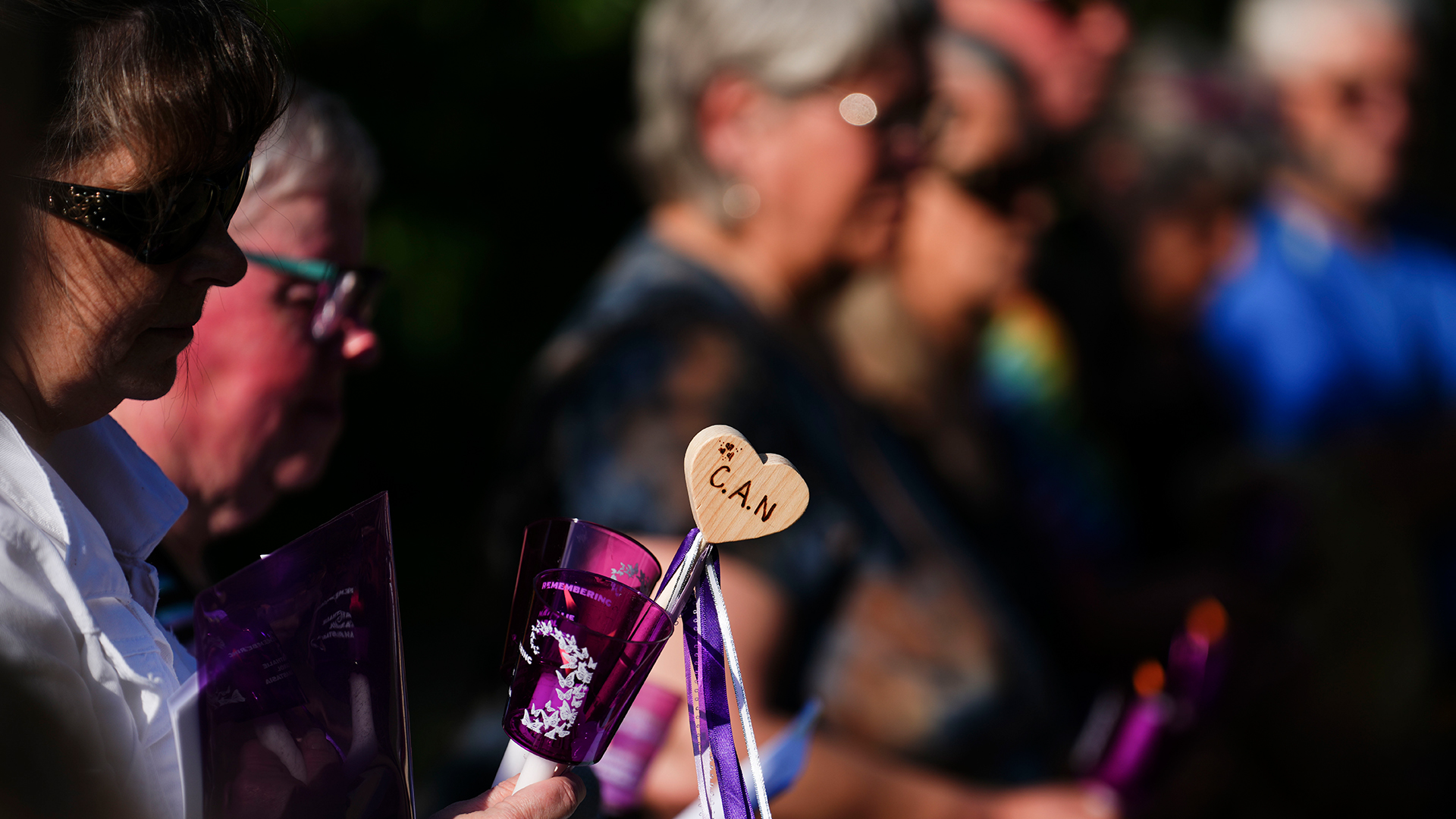 The little heart is fashioned out of plywood and is attached to a long piece of plastic with purple ribbons a couple of inches long hanging from it. The wooden piece would fit in the palm of your hand. The woman holding it has wraparound sunglasses and a white shirt. She’s standing in a row of women who are holding similar items. The mood is somber, with shadows and sunlight intermingling across the faces of the group.