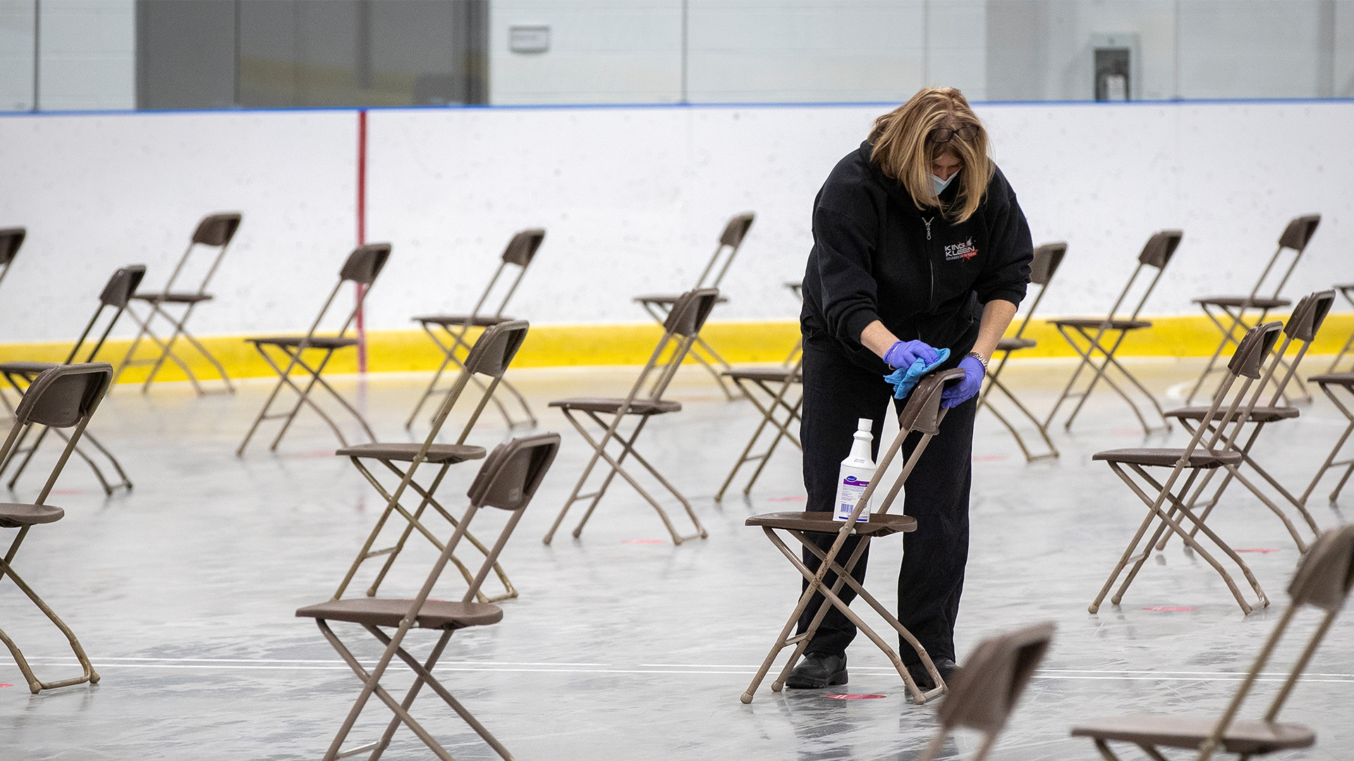 The woman is hunched over with blue cloth in her hand. She is wearing a black sweater, black pants and black shoes, and purple rubber gloves. The chairs are thin, brown and plastic, the kind that fold up. They are placed in rows with lots of space between them. The woman is the only person there. The floor of the rink is light brown.