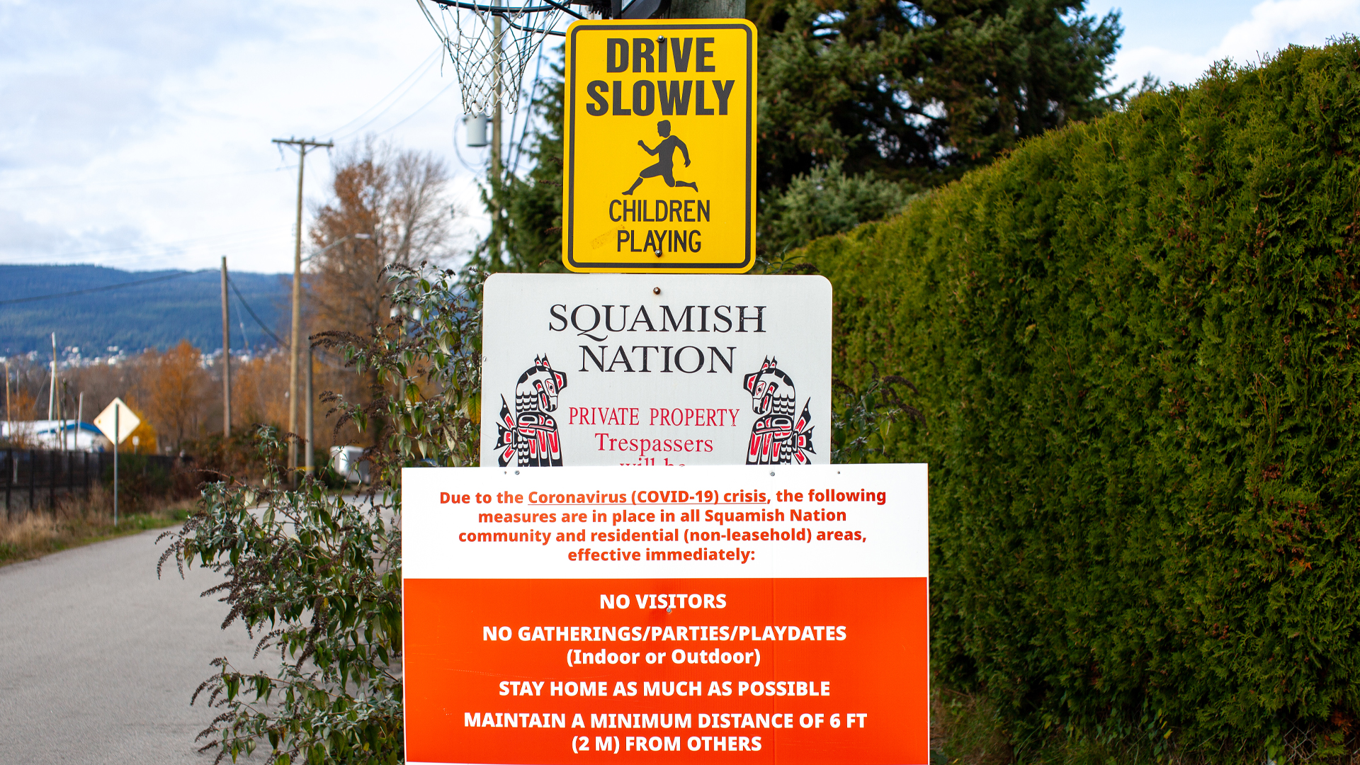 Trois affiches sont visibles sur un poteau qui s’élève en bordure d’une route pavée. Celle du haut invite à circuler à basse vitesse en raison de la présence possible d’enfants, celle du milieu signale que les lieux sont une propriété privée de la nation Squamish et la dernière, plus grande, recommande aux gens de s’isoler chez eux ou, à défaut, de maintenir une distanciation sociale. Un filet de basket-ball est visible derrière les panneaux et l’on aperçoit un village en arrière-plan.