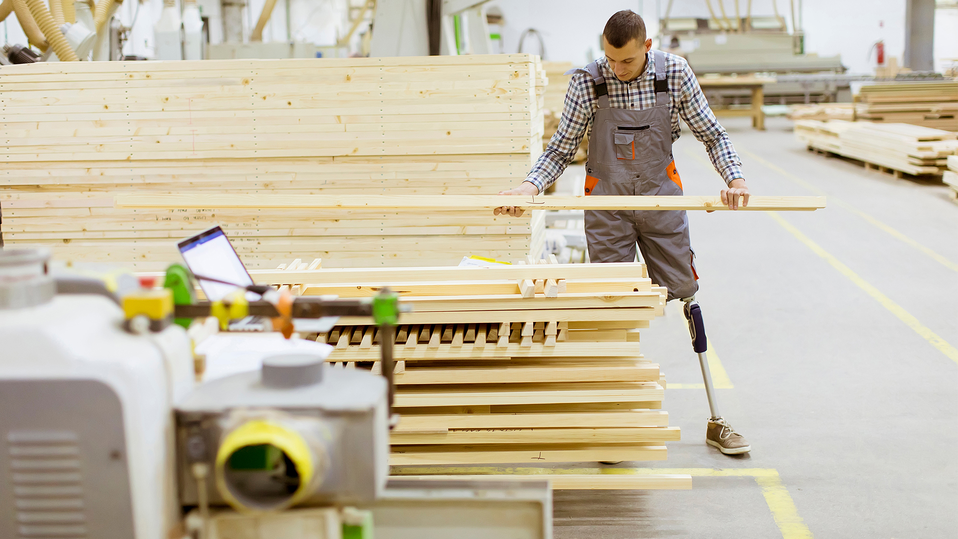 Young man with an artificial leg is working in a furniture factory. He is standing next to a large stack of wood, holding a two-by-four. He is wearing grey coveralls, a plaid shirt and light-brown suede shoes. He uses an artificial limb from the knee down on his left leg. It is a thin metal pole with a slightly larger dark blue piece of plastic just below the knee.