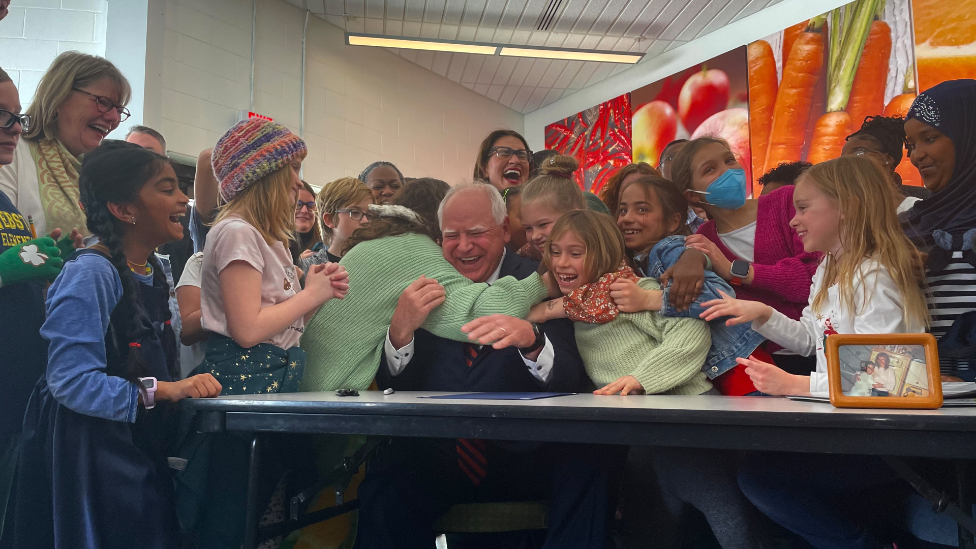 Une vingtaine d’enfants de toutes origines entourent et enlacent le gouverneur du Minnesota, Tim Walz, assis à une table. Quelques adultes sont présents sur la photo. À l’arrière, on peut voir sur un mur de grandes photos de fruits et de légumes.