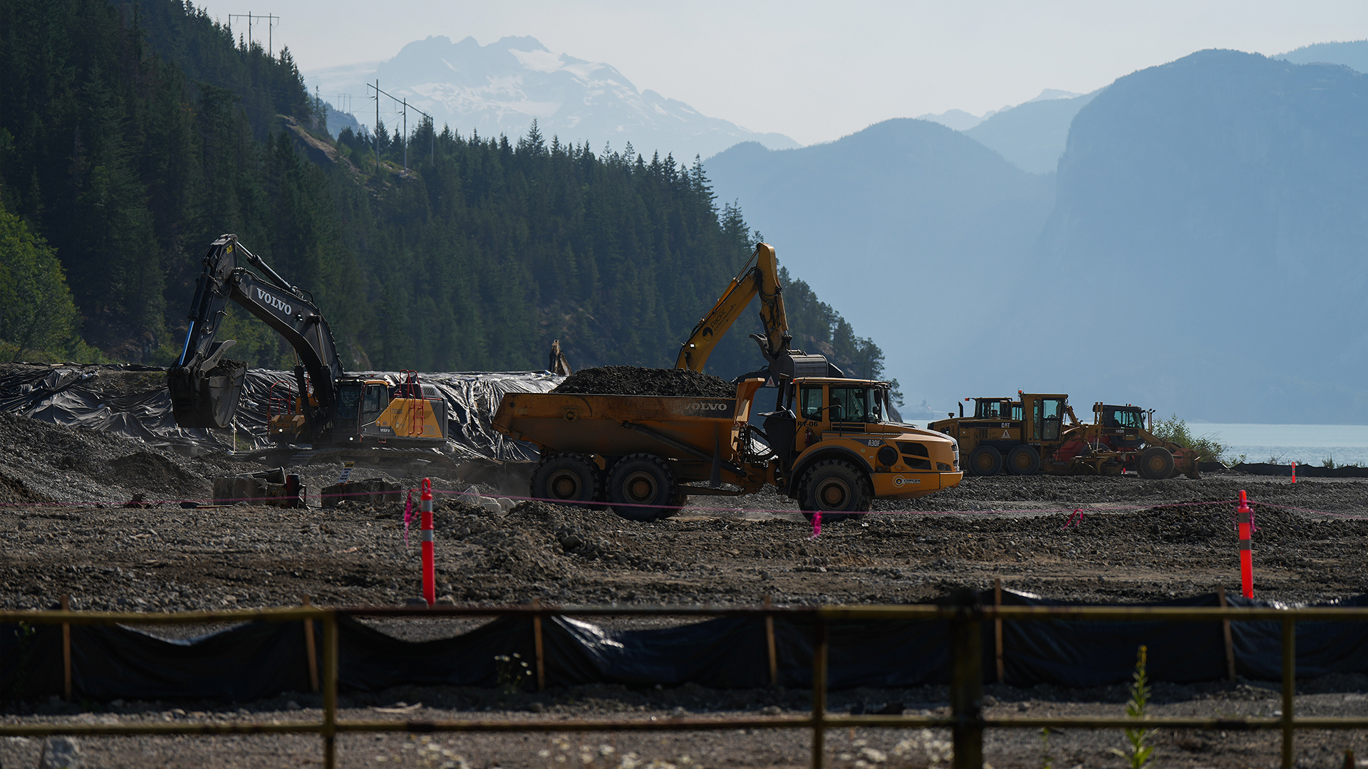 With mountains in the distance, and a forest of tight, tall evergreens in the foreground, two excavators dig a section of flat earth next to the blue-green water nearby. A load of earth fills a dump truck. Three tractors sit a small distance away.