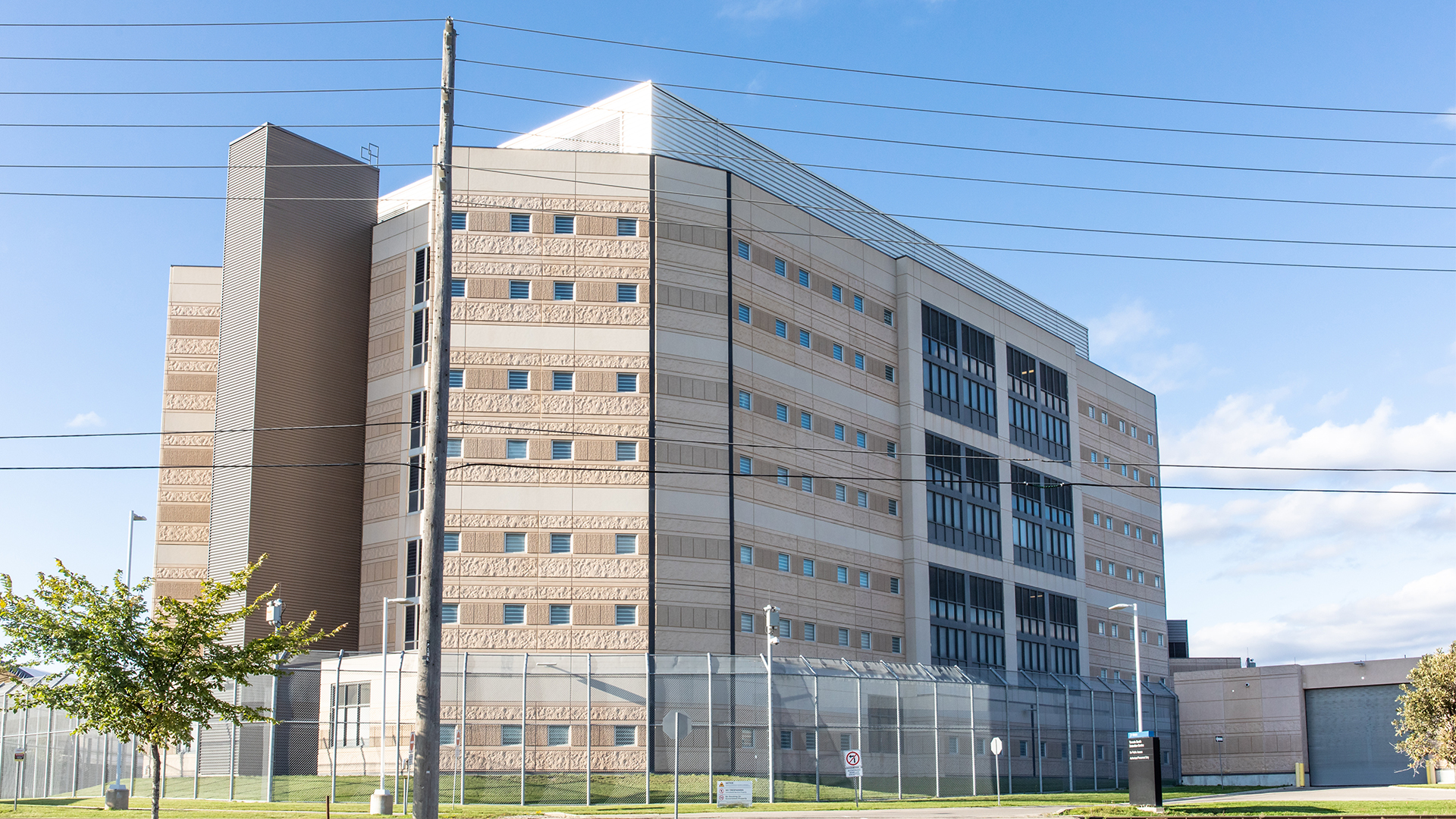 A stone building about eight floors high sits behind a high chain-link fence. The brick and stone is light brown and off-white. The rows of square windows are tiny.
