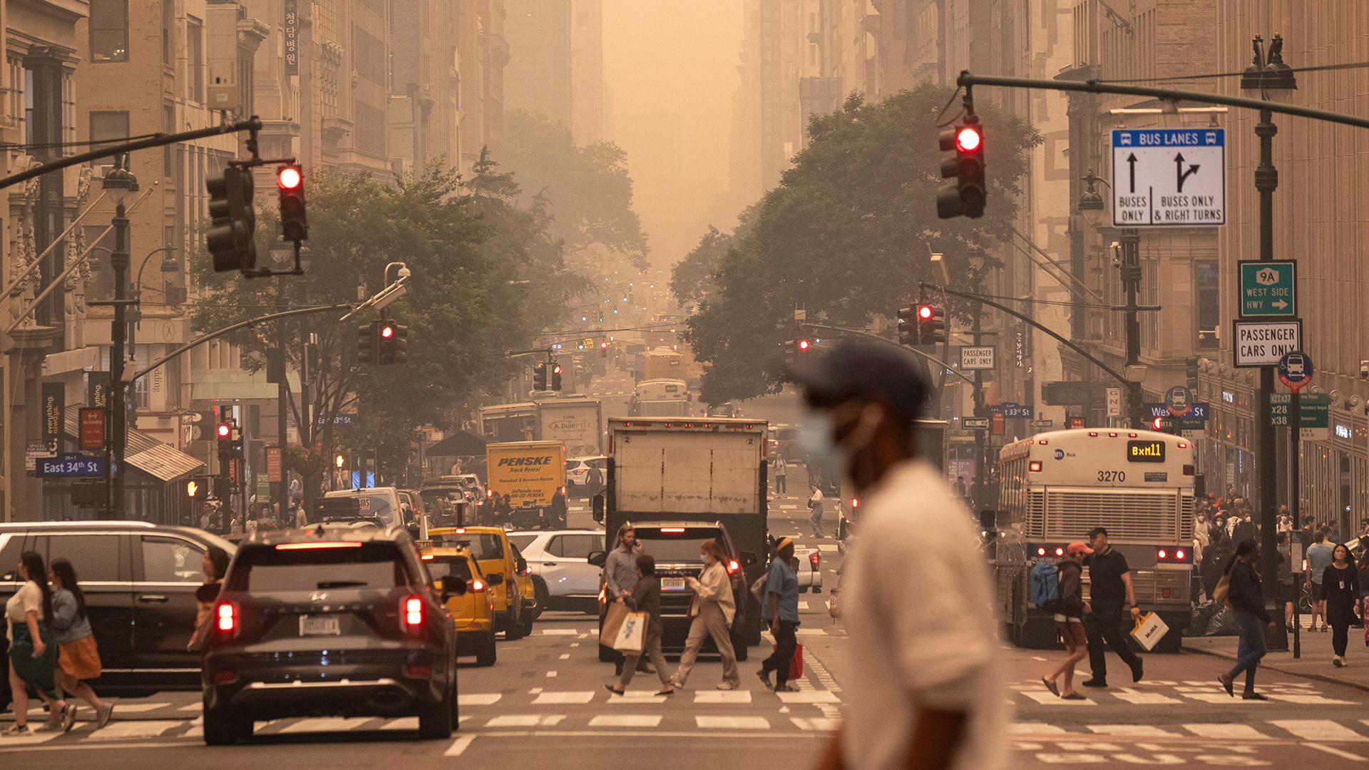 A man wears a mask as he crosses an intersection. The sky is a haze of pale orange, with low visibility a few blocks away. The street is four lanes, filled with cars, trucks and people crossing at the crosswalk.