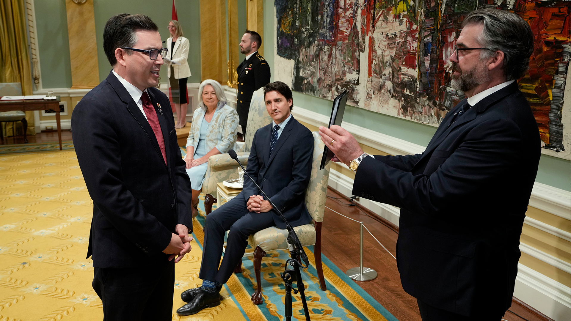 Beech stands and reads text from a tablet held in front of him by a man in a suit. He is in an ornate room with tall ceilings, a golden carpet, green walls with candle sconces and a large piece of art with busy brushstrokes in black, brown, red and creamy white. Seated nearby is the prime minister, Justin Trudeau, and the governor general, Mary Simon, both watching Beech impassively.