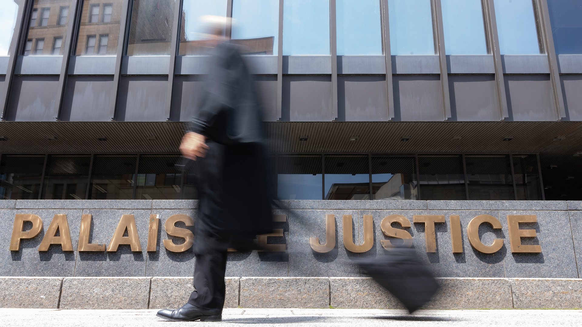 A man in a dark suit with a rolling document case walks by the Montreal courthouse sign that reads ‘Palais de justice.’