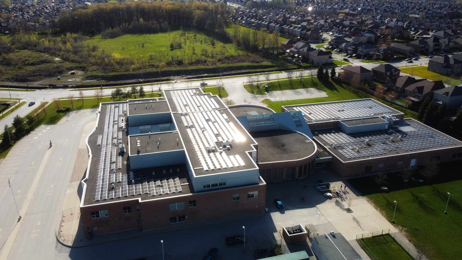 A brick school several stories high has probably close to a hundred panels covering its flat roof. They glisten in the sun. A parking lot surrounds the school, and adjacent to it in the distance is a green field bordered with a small forest and a densely packed suburb of many homes.