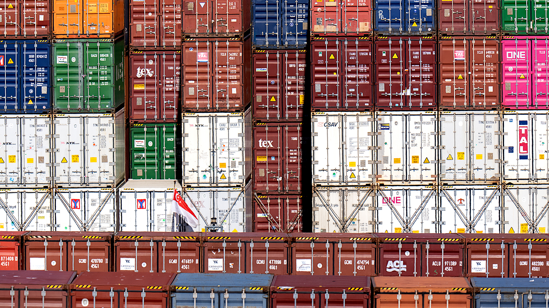 Dozens of containers are stacked on top of each other. The stacks are six high and probably higher. Most containers are rusty-red in colour, and others are white, green, yellow, green, dark blue and grey.