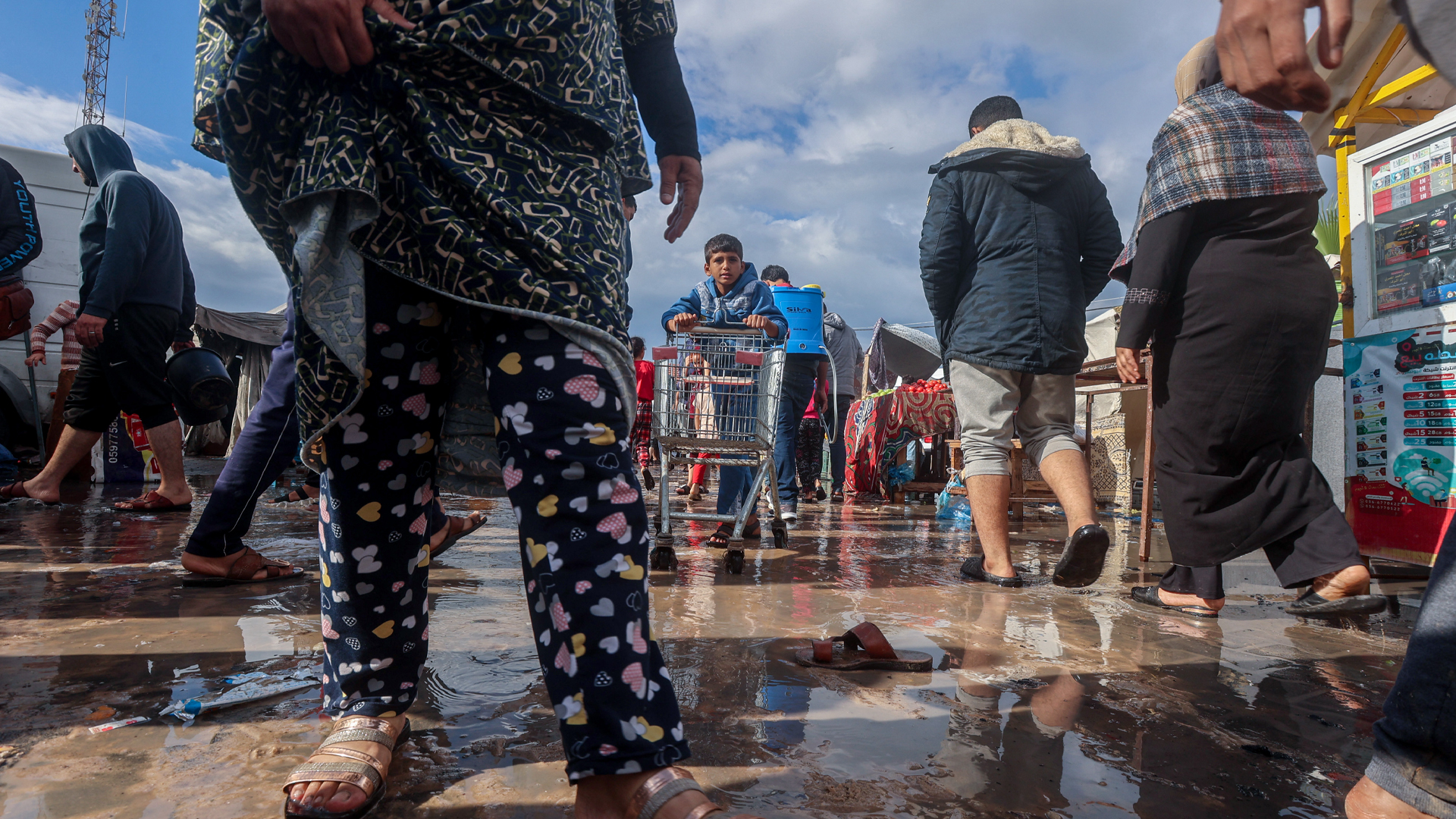 A young boy pushes a shopping cart on muddy ground while adults circulate around him in sandals, holding up pant legs to keep them out of the mud.