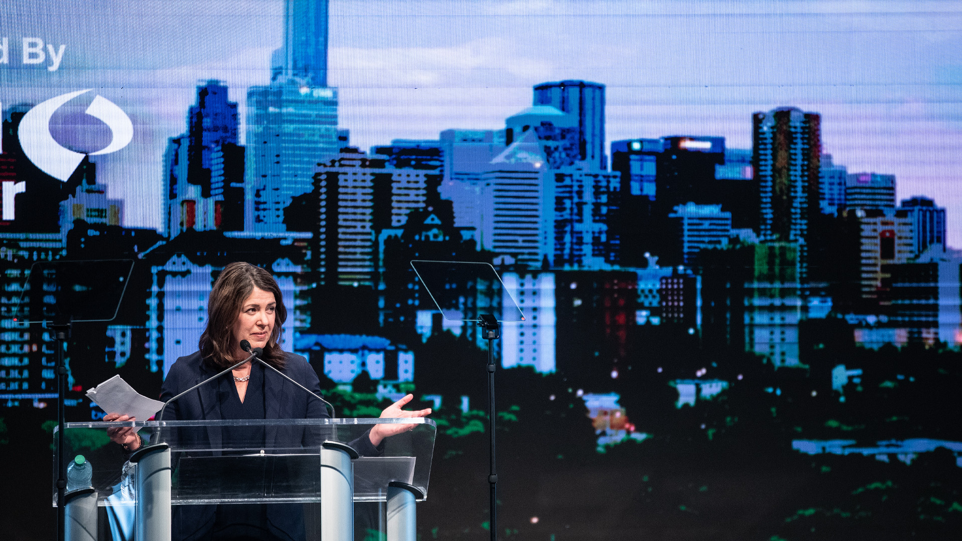 Danielle Smith speaks at a glass podium, speaking notes in one hand, with a stylized image of the Edmonton skyline filling the background.