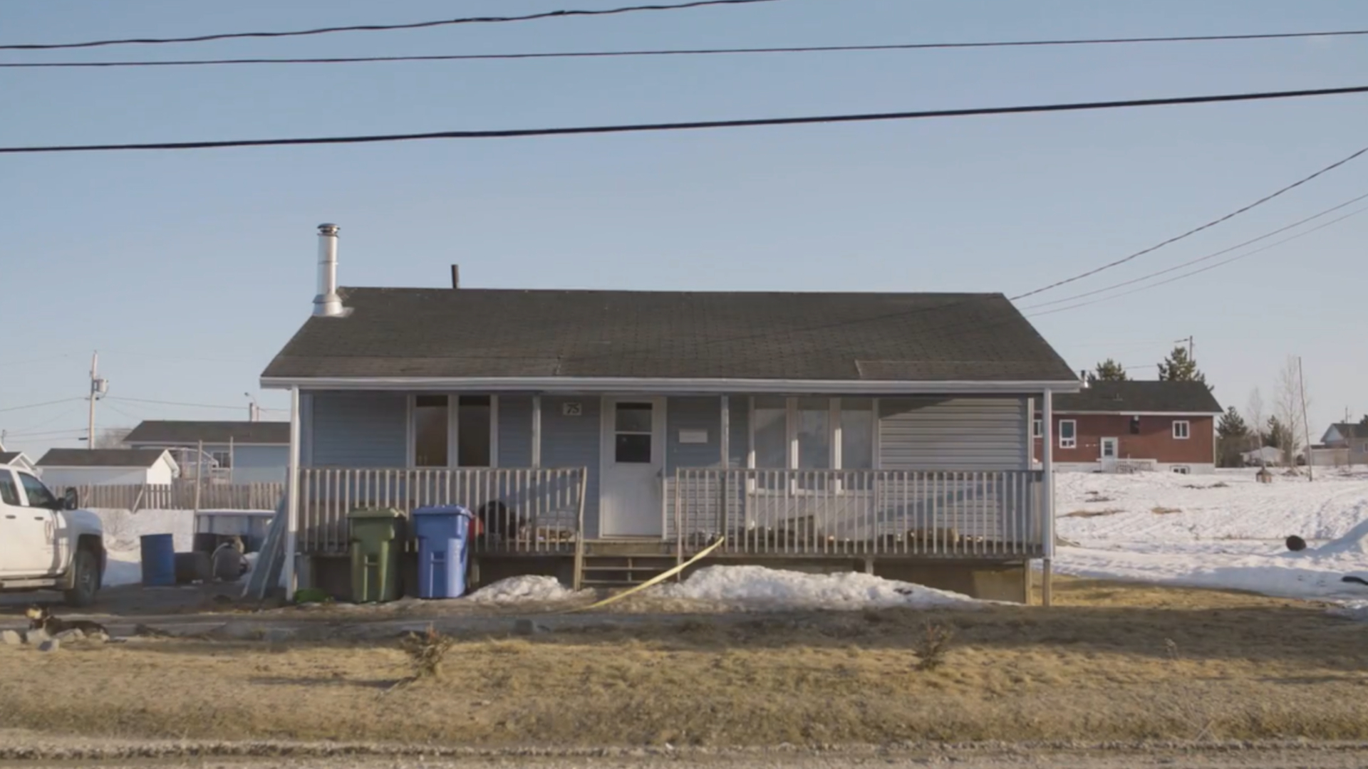 Un petit bungalow gris pâle se trouve non loin de plusieurs autres maisons semblables. Il possède une véranda en bois sur toute sa longueur, une porte d'entrée blanche et deux fenêtres à cadre blanc à l'avant, l'une à trois carreaux, l'autre à deux. Deux poubelles pour les ordures et le recyclage sont adossées à la véranda. Une camionnette blanche est garée dans l'allée. Des restes de neige se trouvent dans la cour d'entrée.