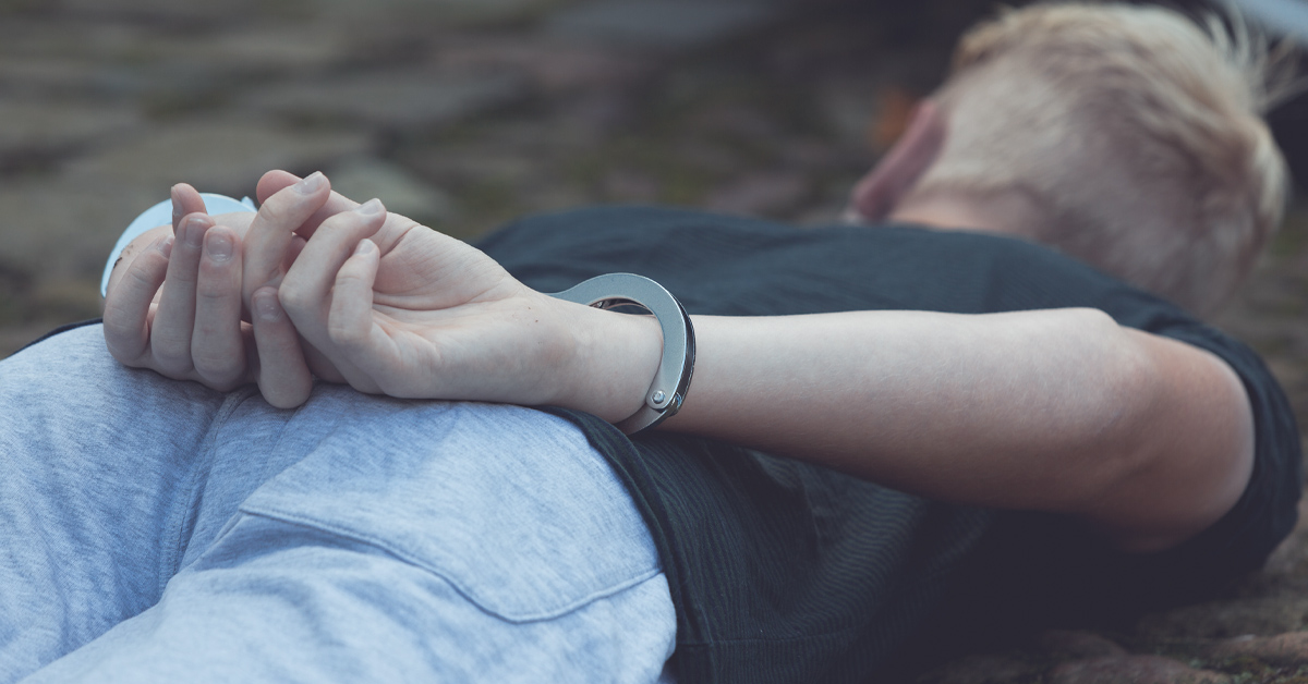 A teenage boy has his hands cuffed behind his back while lying on the ground near a car.