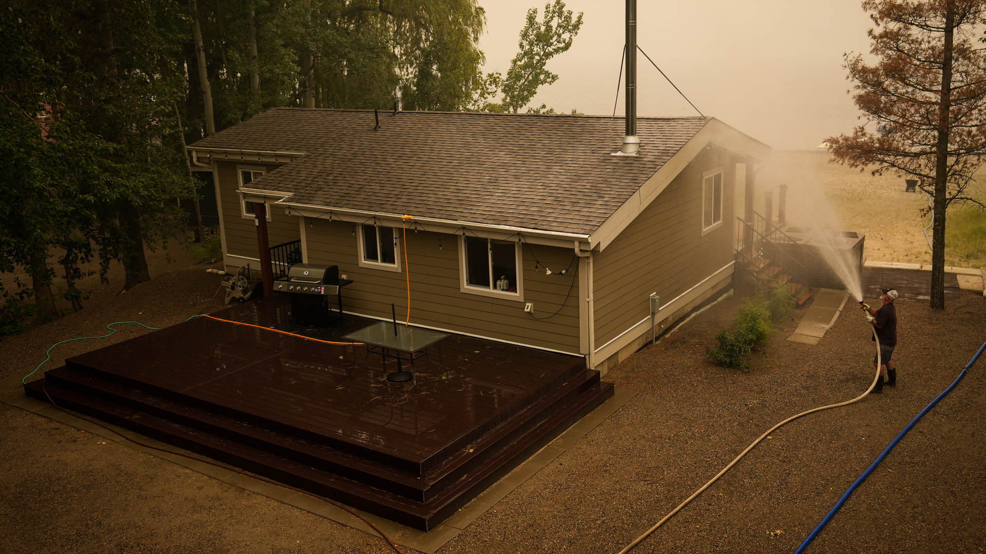 The man is standing a few feet from his bungalow with water blasting from a hose in his hands. He is aiming the water spray at the roof. The house is surrounded by a dense stand of tall trees on one side. In the distance, smoke fills the air, which has a warm-coloured haze.