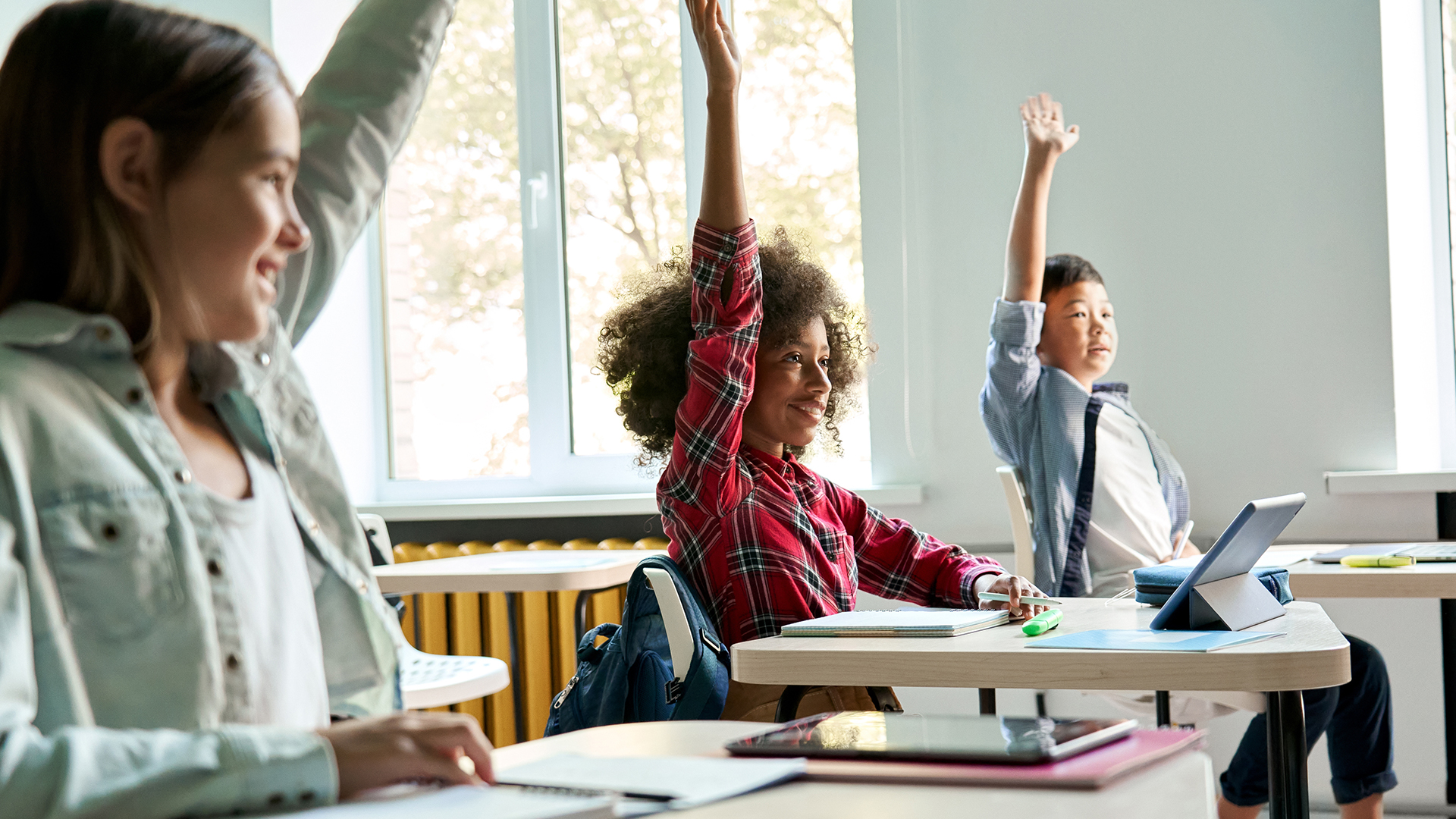Three elementary-age girls sitting at desks are smiling with their hands up to ask a question. The room is bright with tall windows.