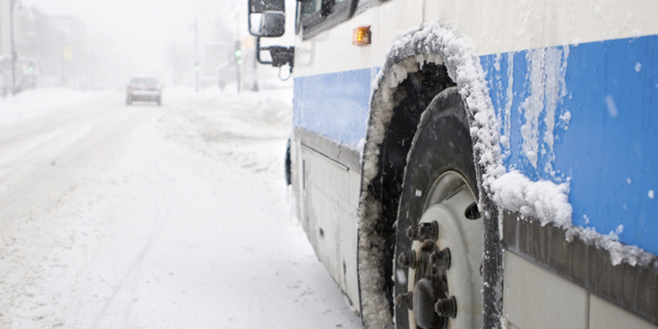 View of the bus from just behind the front left wheel. The rim is covered in slush, and pieces of ice have formed as rivulets of water trickled down the side of the vehicle, which is white with a blue stripe. The road is covered in snow, and snow is falling.