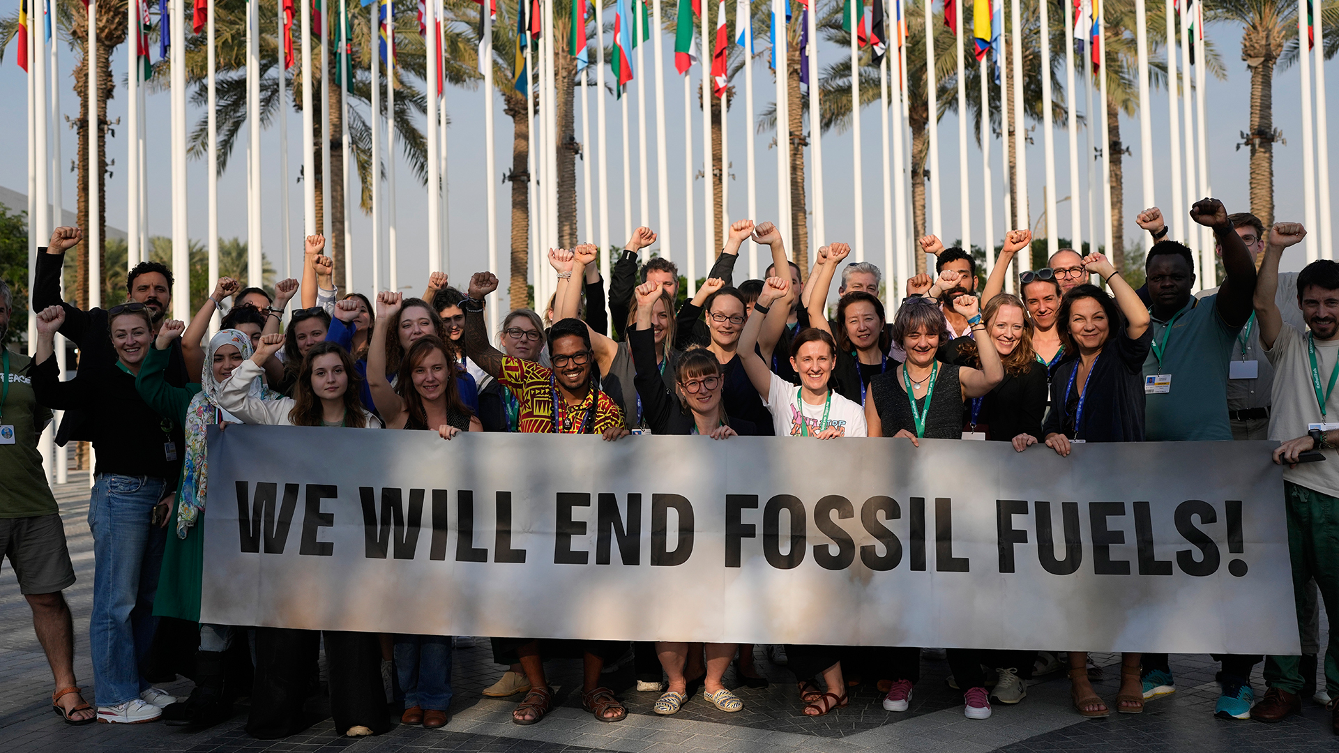 Environ 35 personnes se tiennent derrière une longue pancarte qu'elles tiennent à la main. La plupart d'entre elles ont le poing levé et toutes sourient. Derrière eux se trouve une collection de mâts de drapeaux étroitement espacés, portant les drapeaux colorés de nombreux pays. Tous pendent mollement. Derrière les drapeaux se trouve une rangée de palmiers.