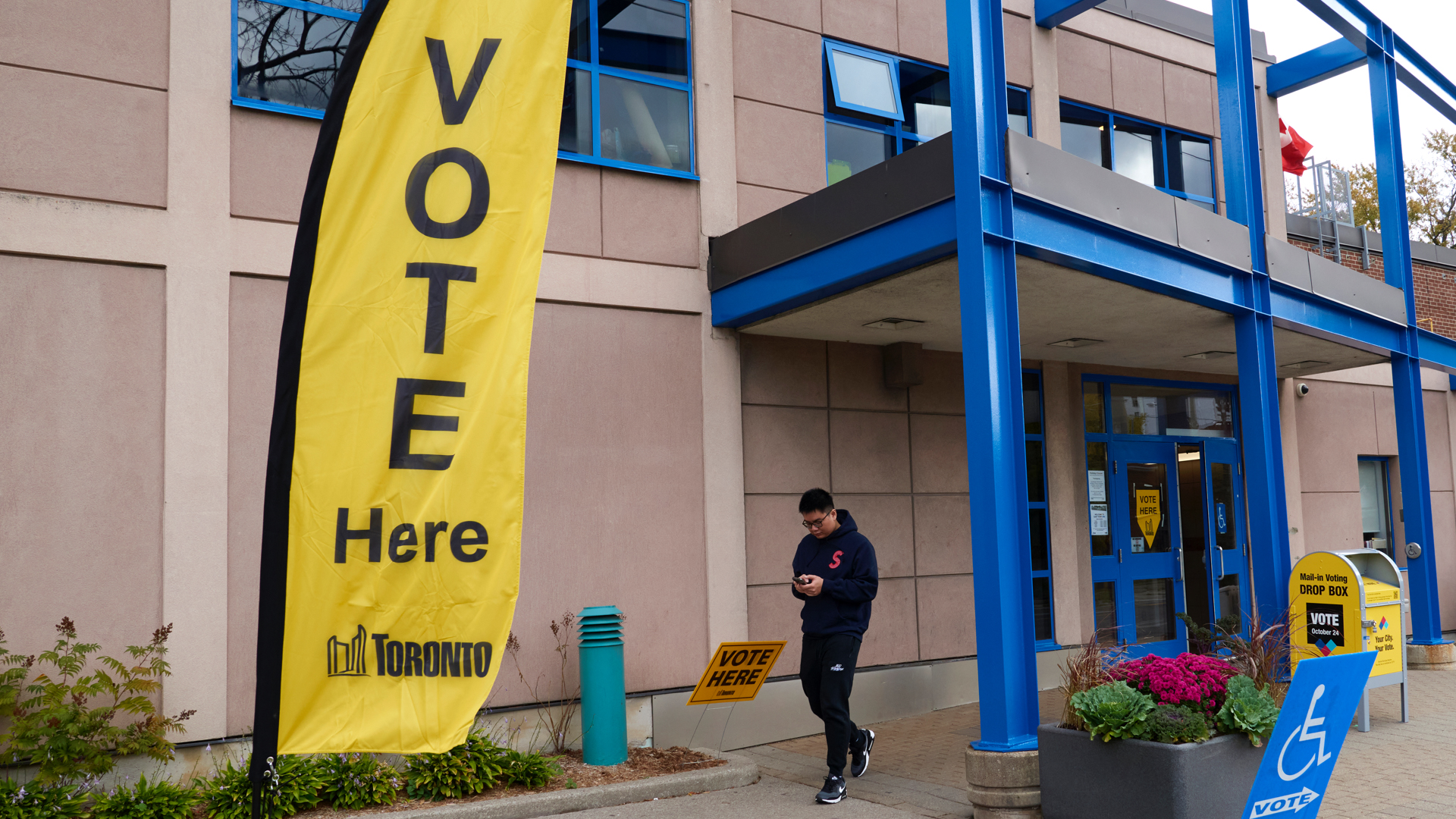 A dark-haired man in a black hoodie, black pants and black sneakers looks at his phone as he leaves the entrance to the centre. The building is two storeys, has bright blue girders that hold a roof over the front doors, which are also bright blue. A tall yellow fabric sign says “Vote here” with a City of Toronto logo at the bottom.