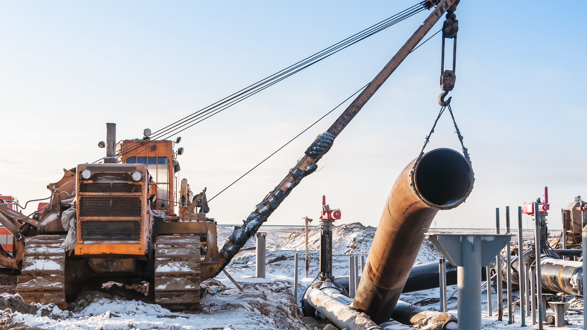 A length of pipe hovers over snow-covered ground on the Prairie, held up by a strap held by a crane lift.