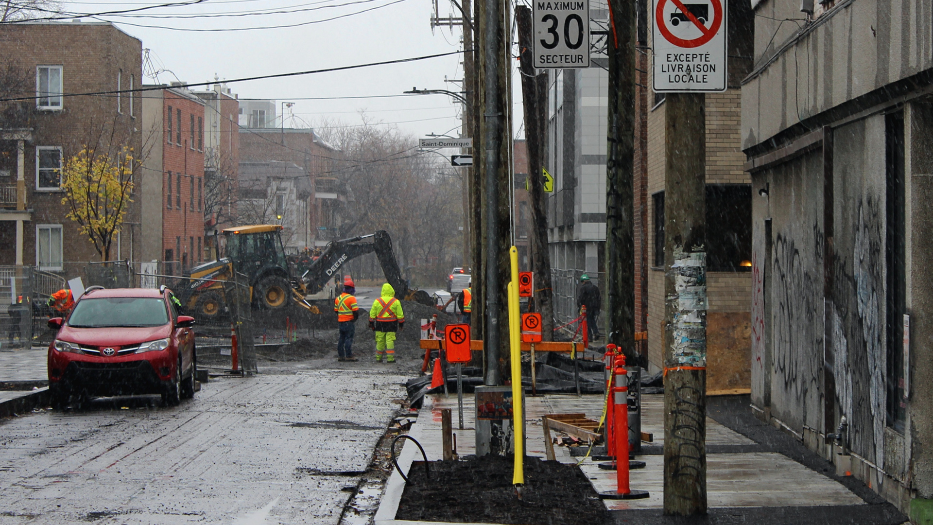 Une excavatrice creuse l’asphalte d’une rue résidentielle dont la surface a été grattée. Une équipe d’environ quatre hommes vêtus de gilets et de pantalons de travail vert et orange fluorescents observe la scène.