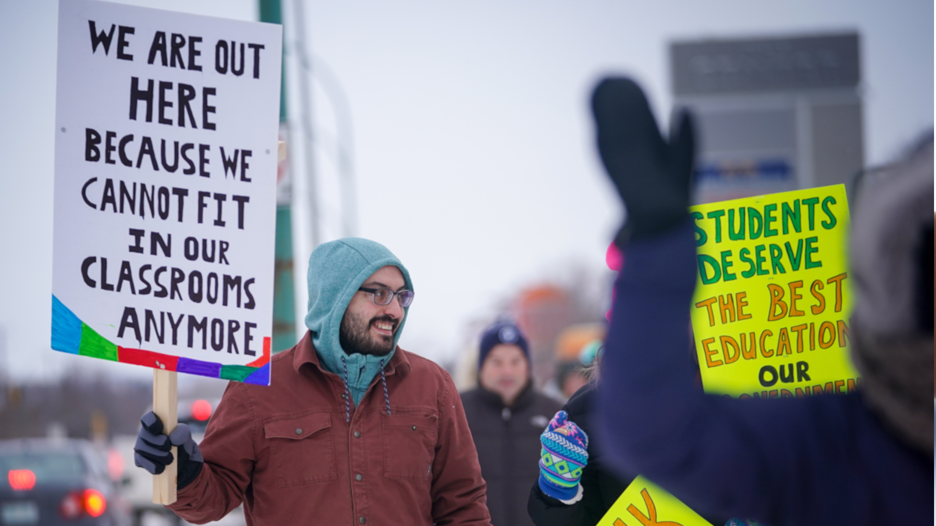 The man is walking with several other people, all holding homemade signs that say “Students deserve the best education our…” (the rest is obscured) and “Honk to support…” and “We are out here because we cannot fit in our classrooms anymore.”
