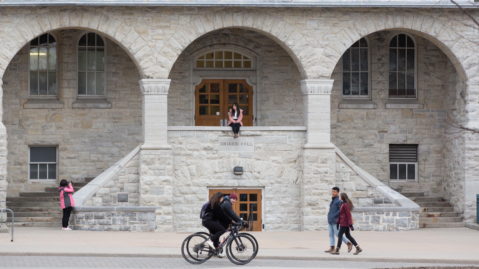 A grey stone university building with young people cycling and walking past while one person in a pink coat sits in an archway posing for a photo.
