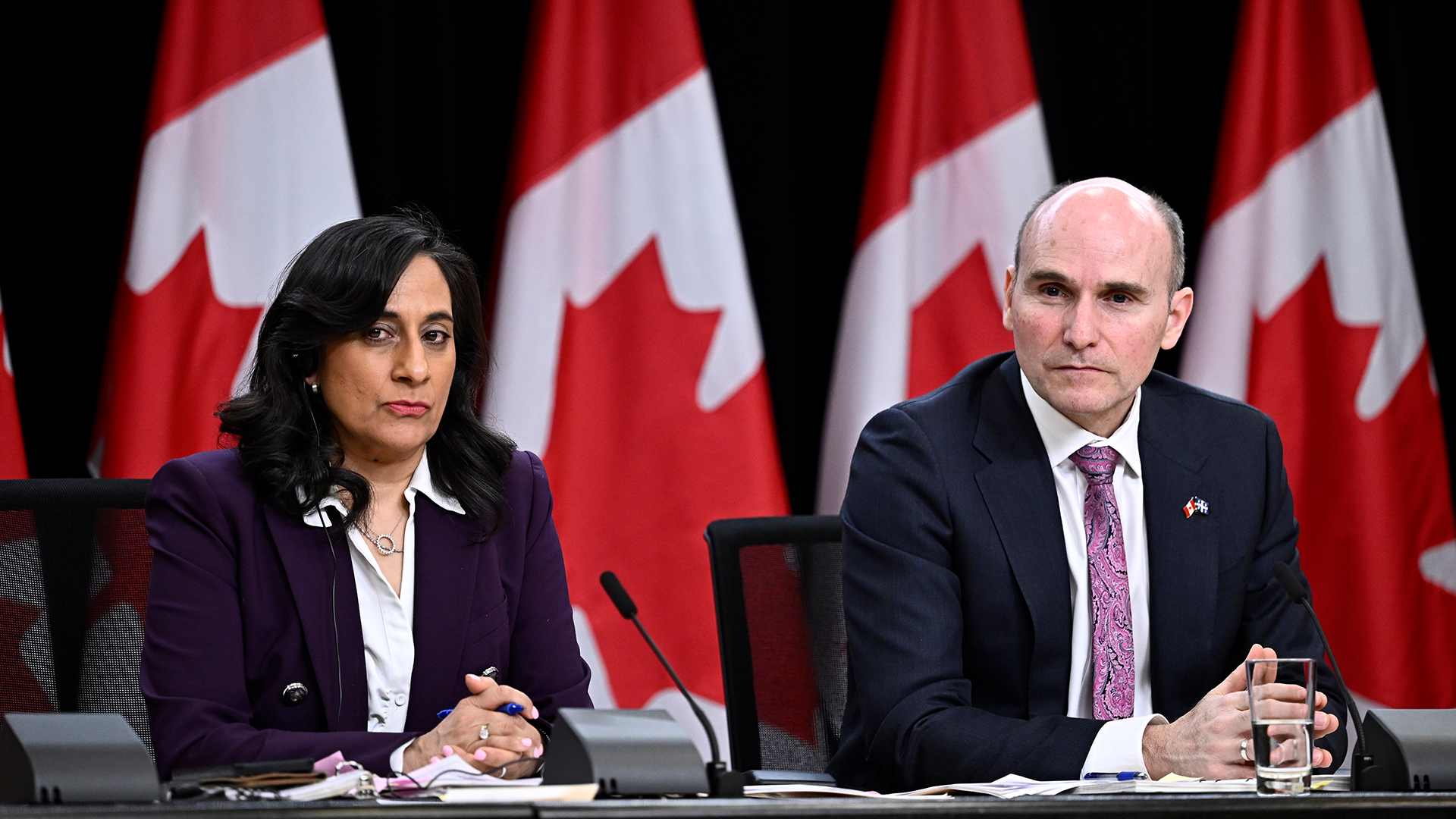 Le ministre des Services publics et de l’Approvisionnement, Jean-Yves Duclos, à droite, et la présidente du Conseil du Trésor, Anita Anand sont assis à un long bureau où se trouve une rangée de drapeaux canadiens Ils sont tous deux vêtus d'un costume sombre et d’une chemise blanche. Ils ont les mains jointes devant eux et s’appuient sur le bureau.