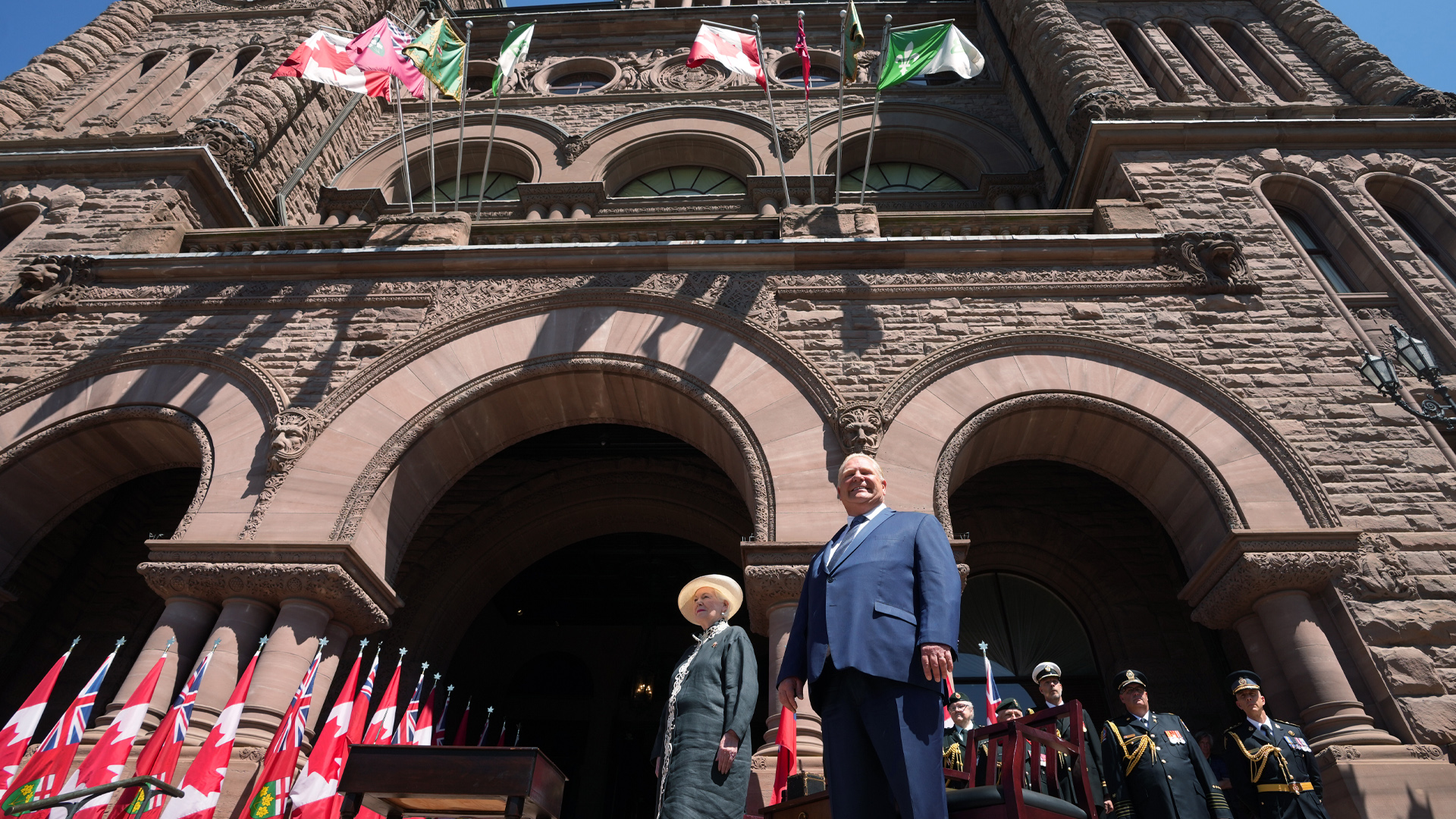 Queen’s Park stands against the blue sky with the lieutenant governor and the premier in the foreground.