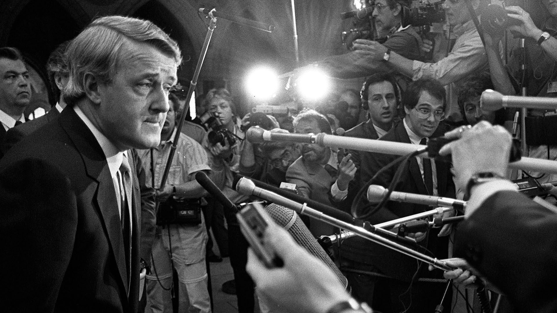 Mulroney is surrounded by a wall of reporters and camera operators. At least nine microphones are pointed at him.